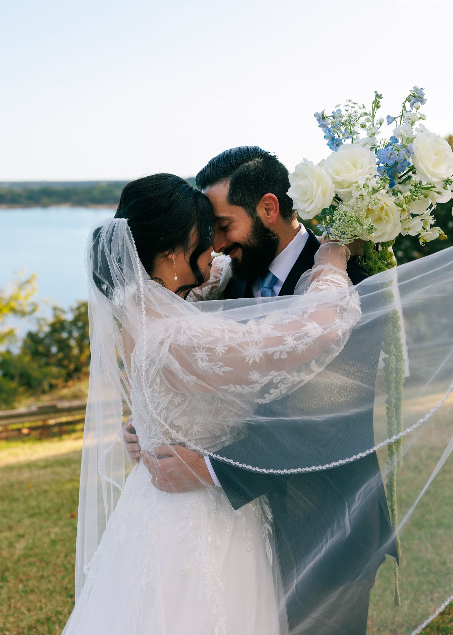 Romantic wedding moment captured against a lakeside backdrop with flowing veil and white bouquet.