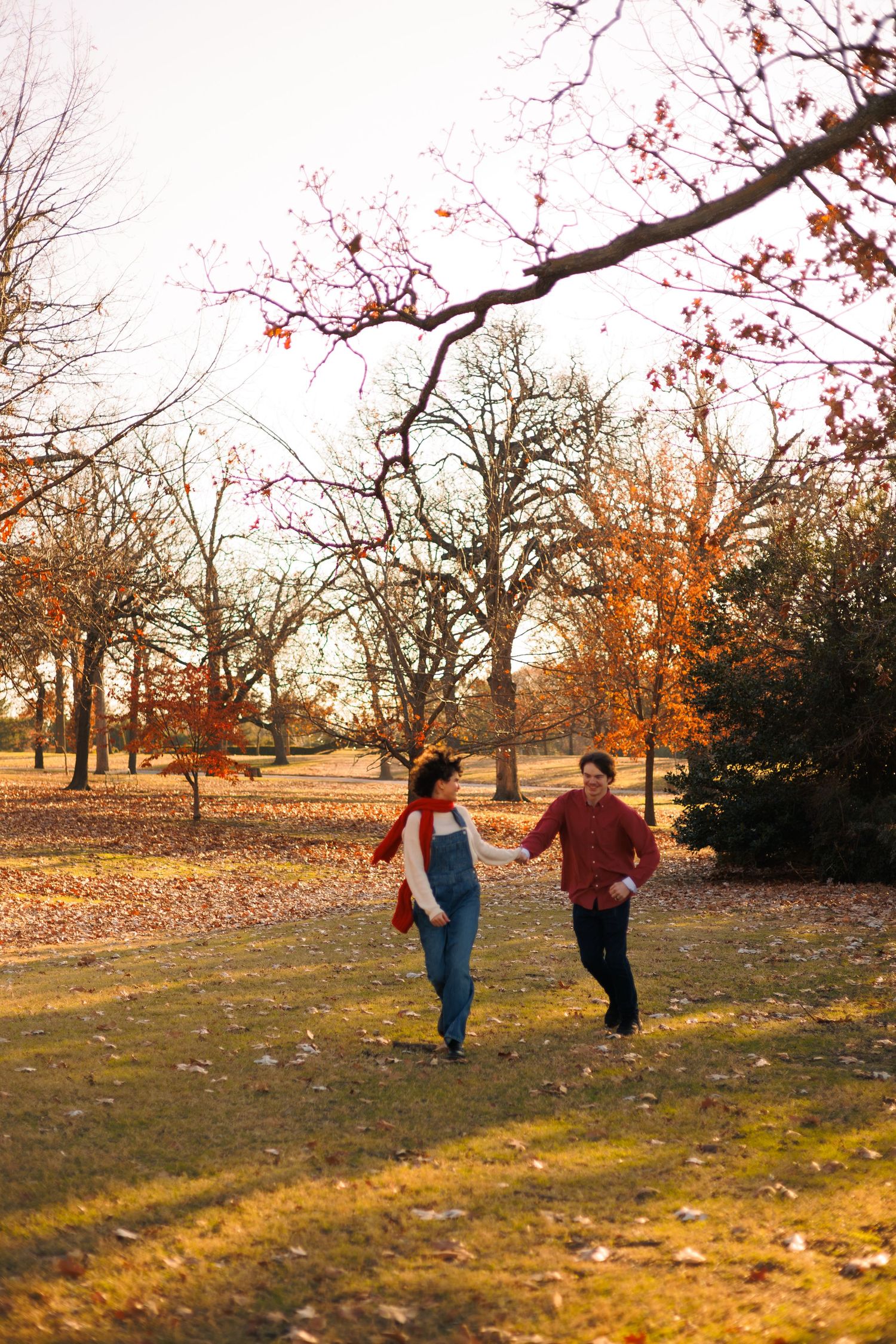 A couple playfully running through a park during golden hour with autumn trees in the background.