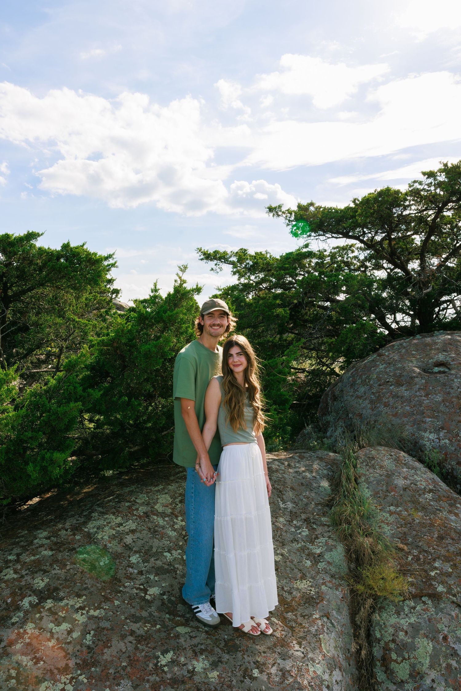 Two people share a tender moment on rocky cliff with green trees in background.