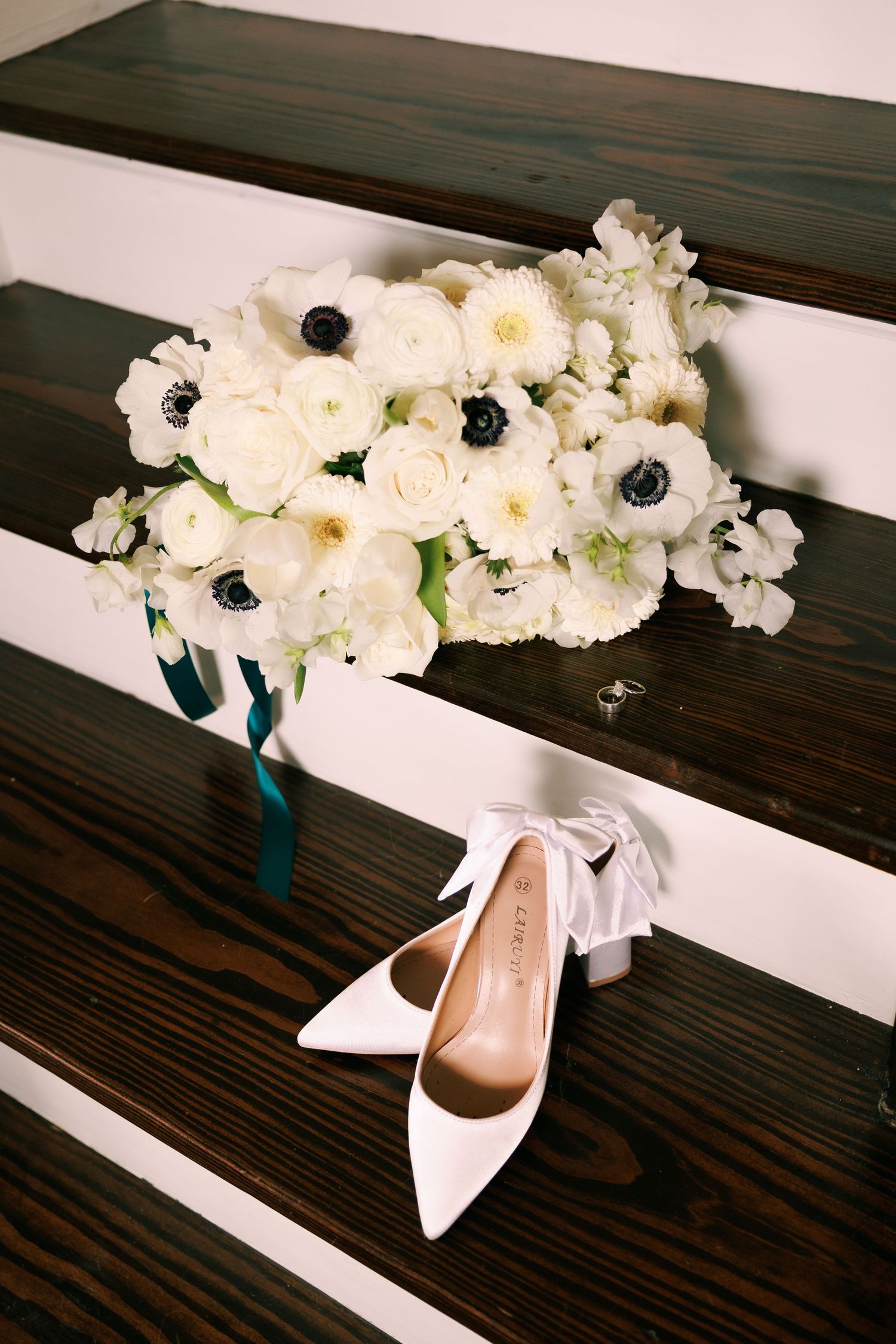 White floral arrangement with bridal shoes displayed on dark wooden bench.