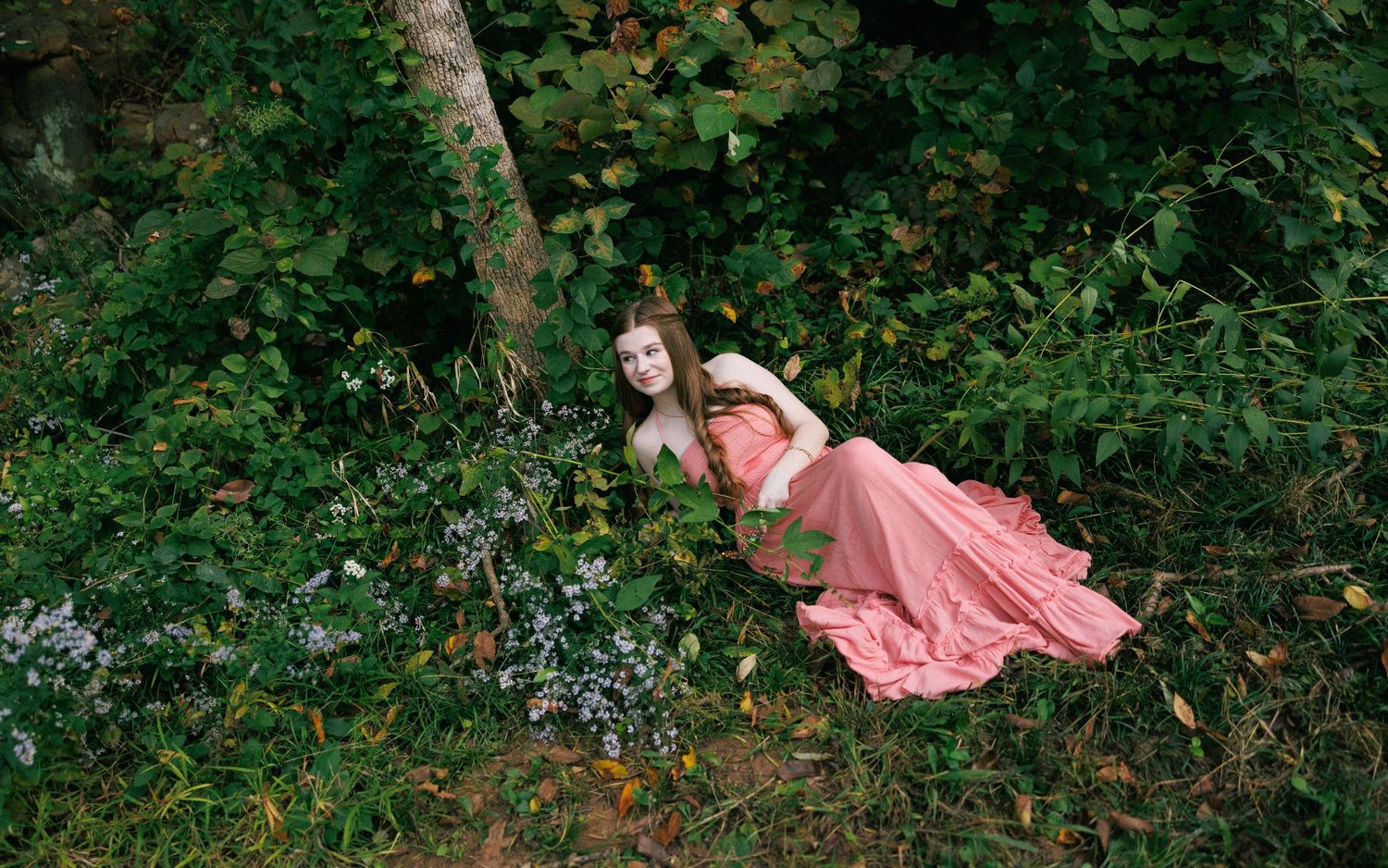 A person in a coral pink dress lies dreamily among lush greenery and wildflowers in a forest setting.