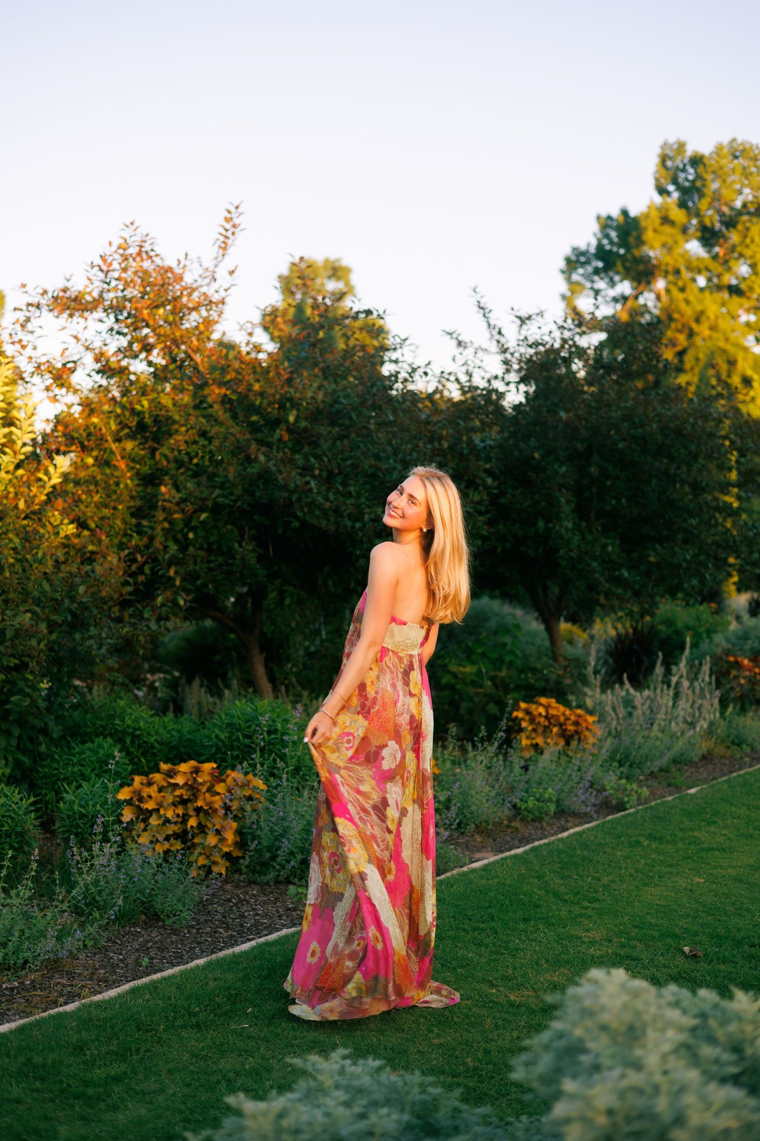 Blonde woman in floral maxi dress posing in lush garden at golden hour sunset.