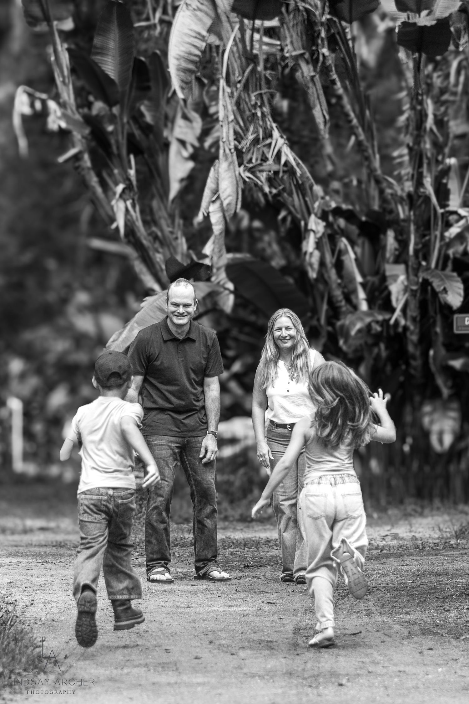 Black and white photo of a family with two children running toward smiling parents on a tree-lined path.
