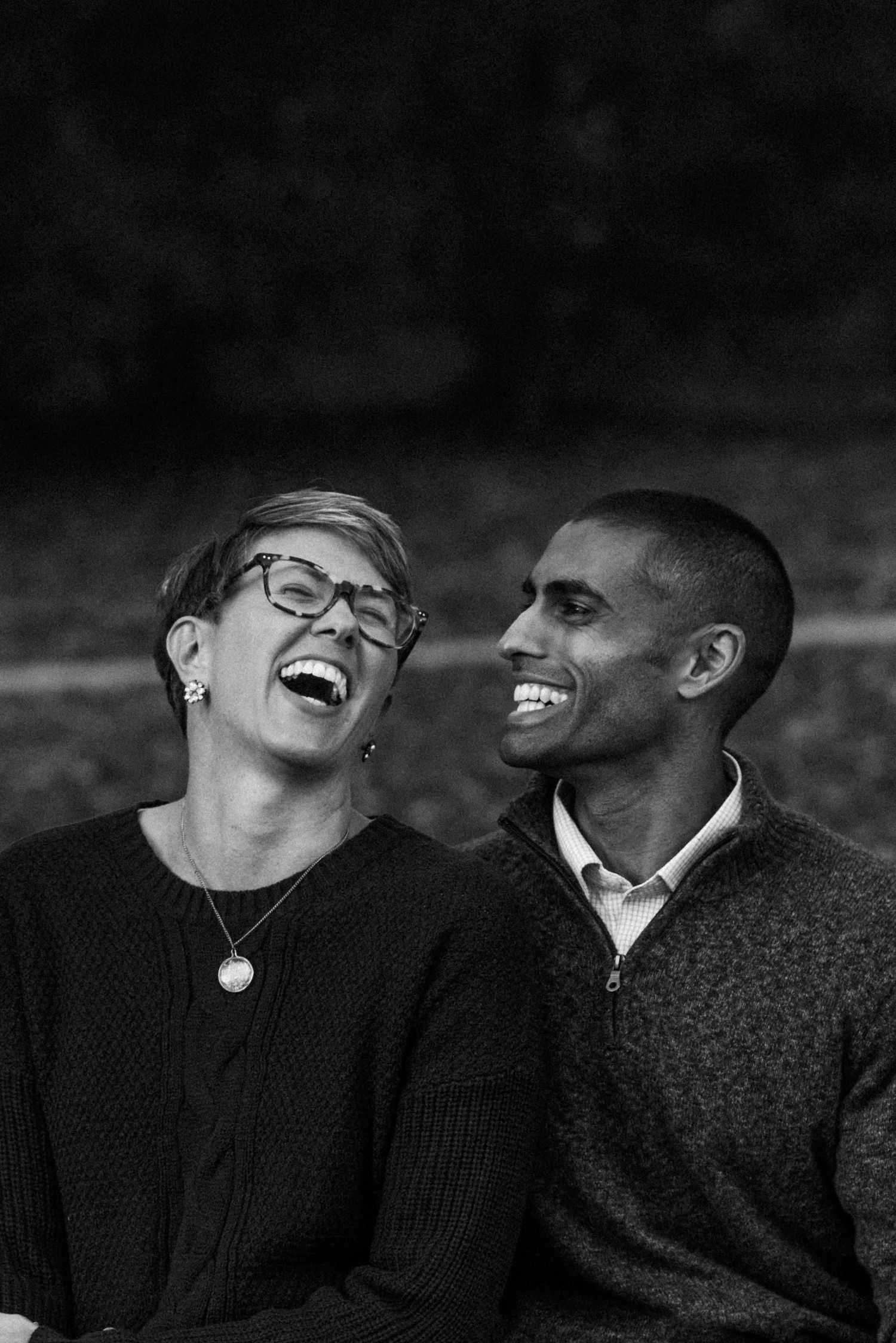 Black and white photo of a laughing couple sitting outdoors, woman with glasses and short hair, man in sweater smiling.