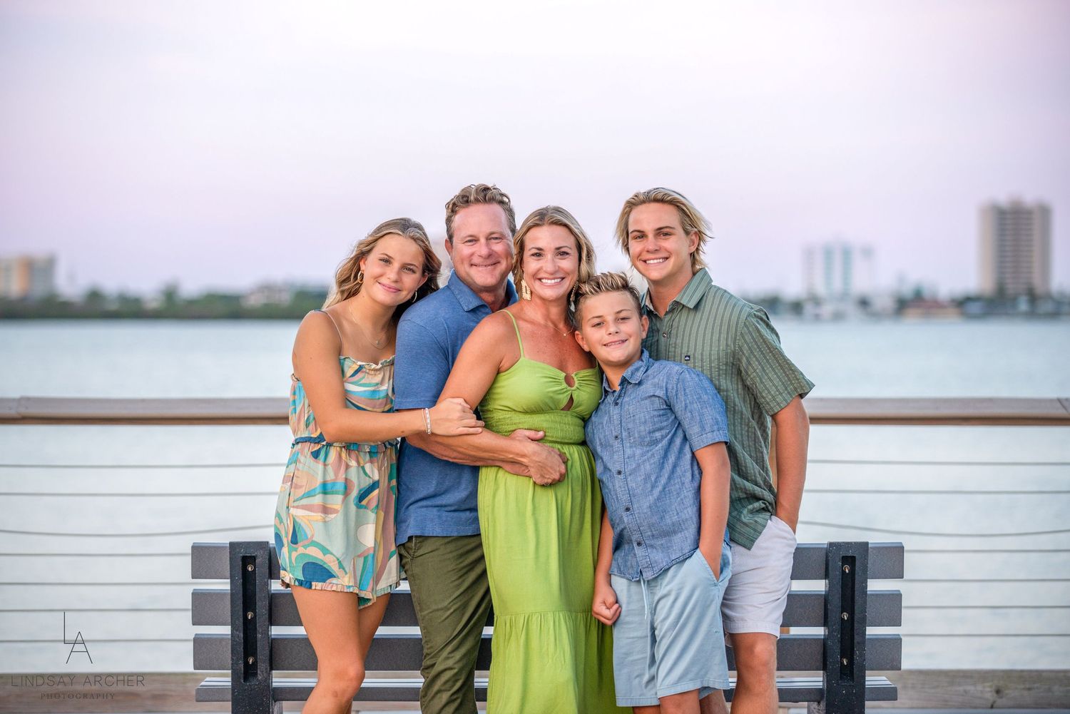 Family of five posing together on a waterfront pier at sunset, smiling warmly in colorful summer outfits.
