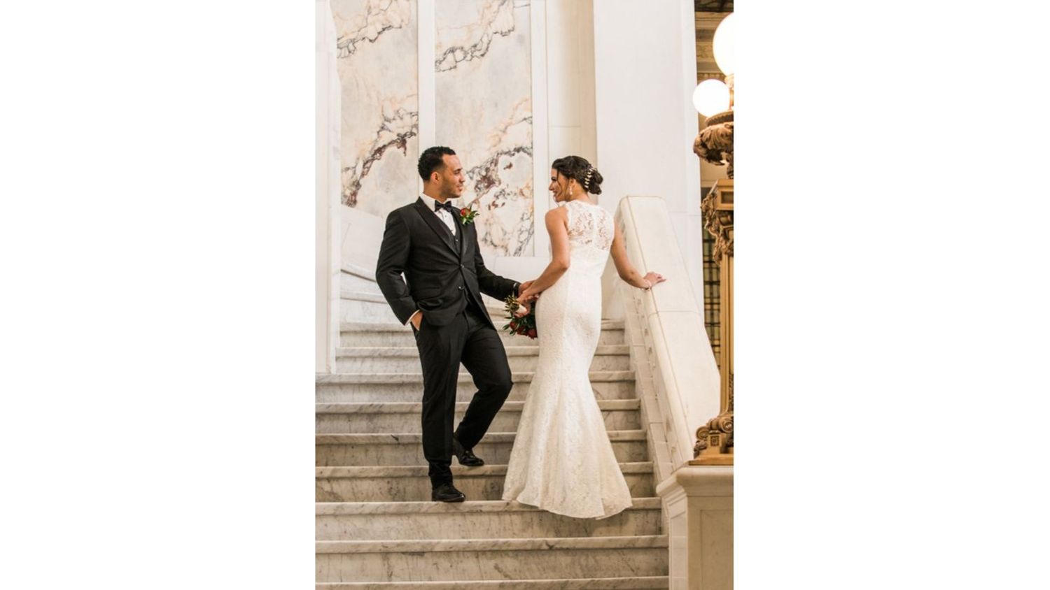 Bride and groom share a romantic moment on grand marble staircase in elegant wedding attire.