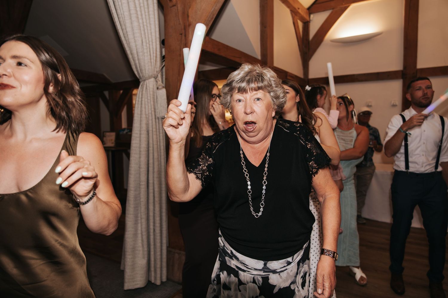 Elderly woman dancing joyfully at a wedding reception, holding a glow stick, surrounded by guests in a rustic venue.