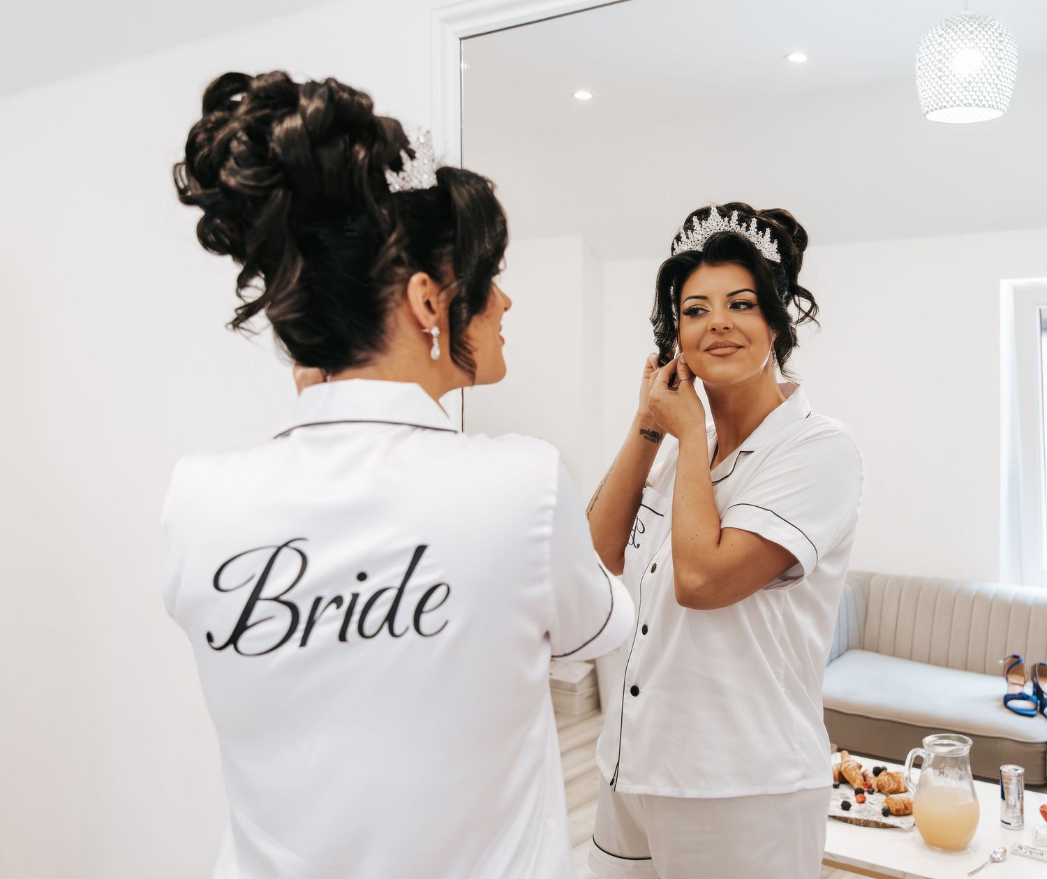 Bride in white robe adjusting tiara while looking in mirror during wedding preparation.