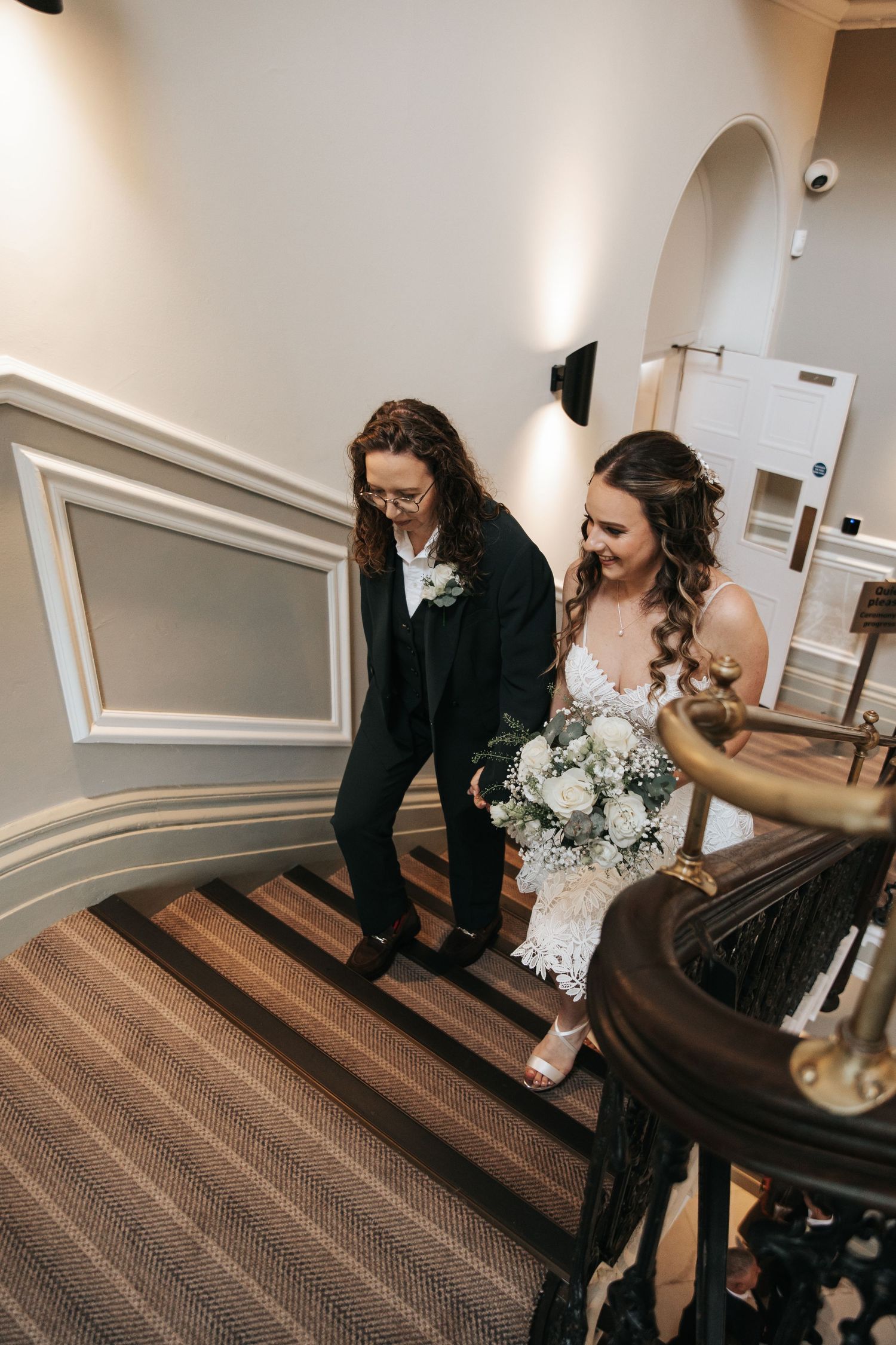 Bride holding white bouquet walks down elegant staircase with partner in black suit at wedding venue.