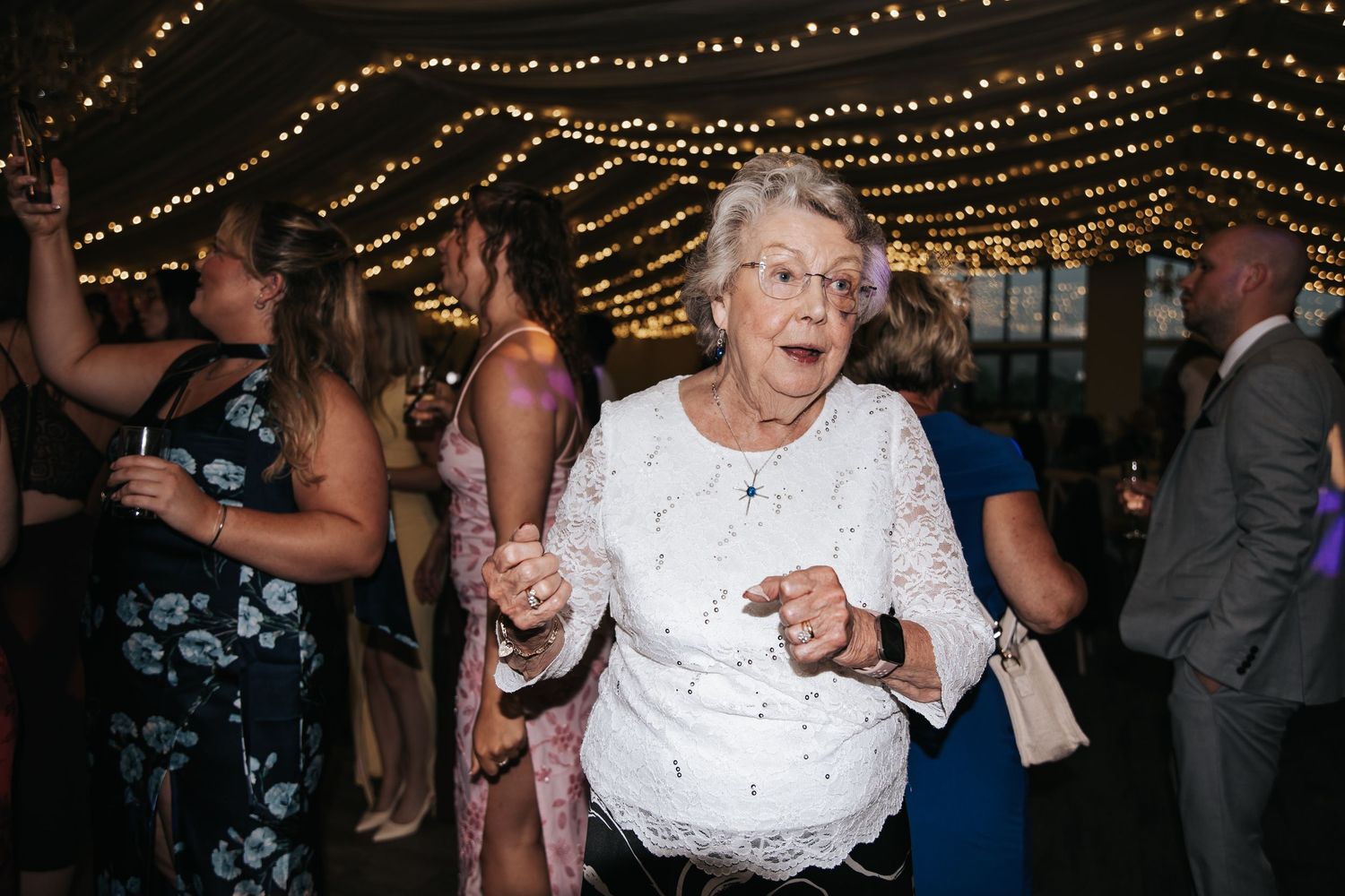 Elderly woman in white lace dress dancing joyfully at a wedding reception under warm string lights.