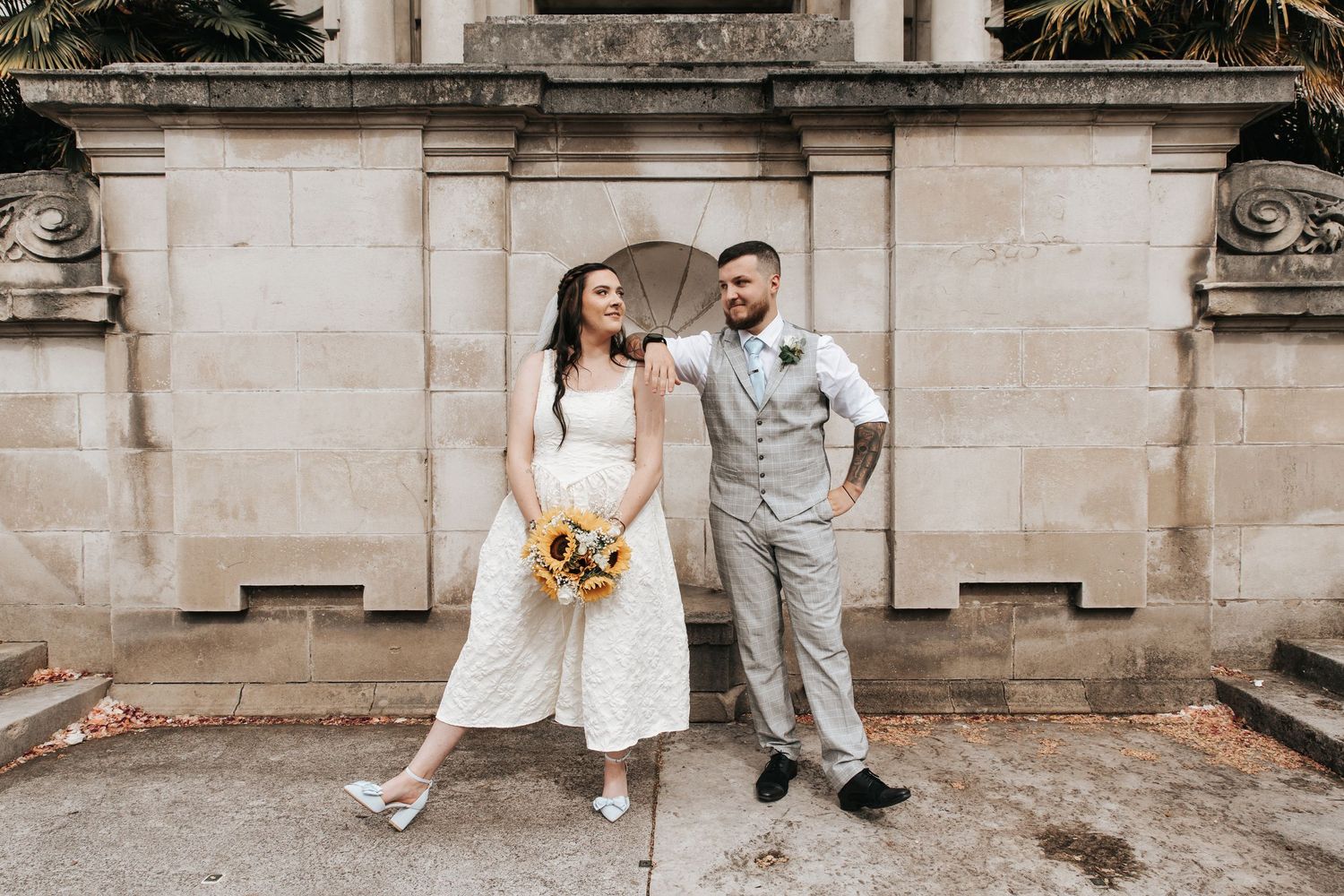 Bride and groom posing together outside a stone building, bride holding sunflower bouquet, groom in gray suit.