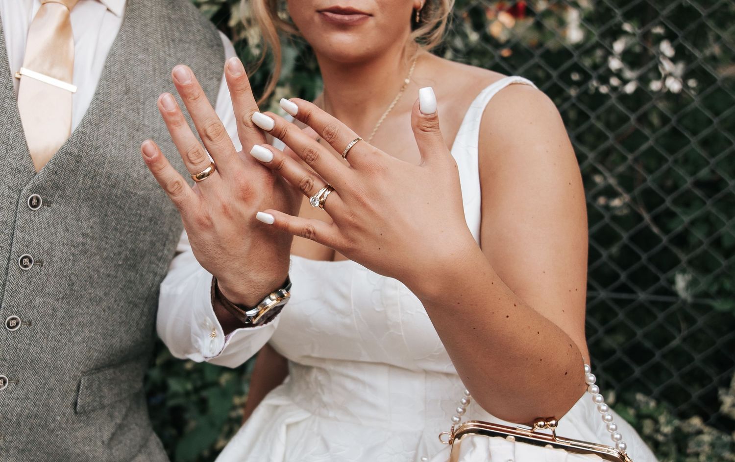 Bride and groom showing off wedding rings, bride wearing white dress with elegant nails, groom in grey suit.