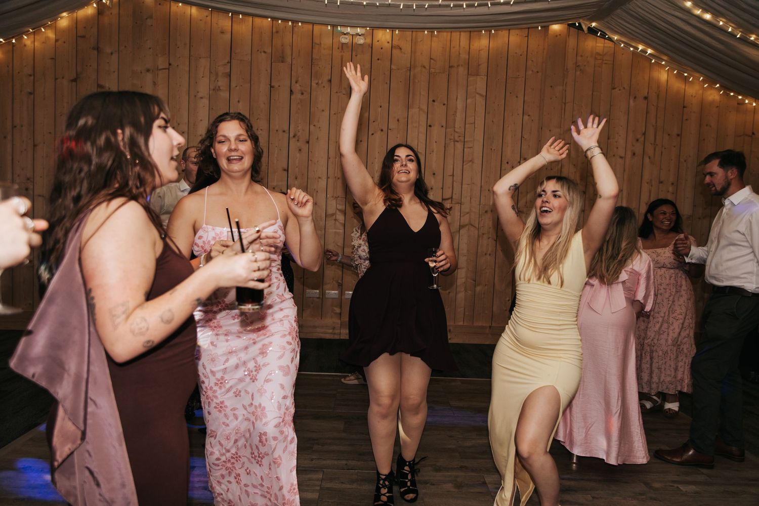 Women dancing joyfully at a wedding reception in a rustic barn venue with string lights.