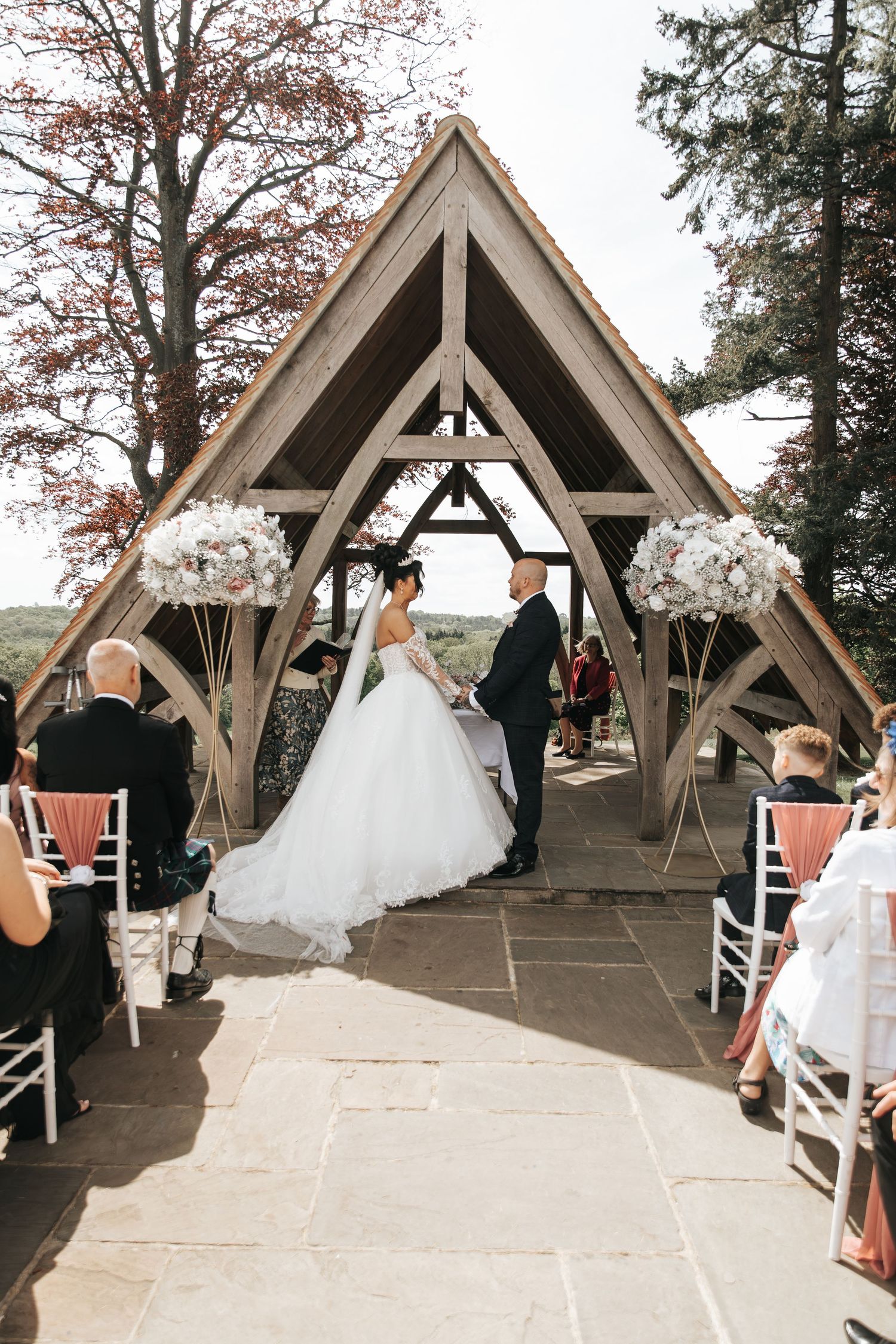 Bride and groom exchange vows under a rustic wooden arch chapel outdoors, flanked by white floral arrangements and seated guests.
