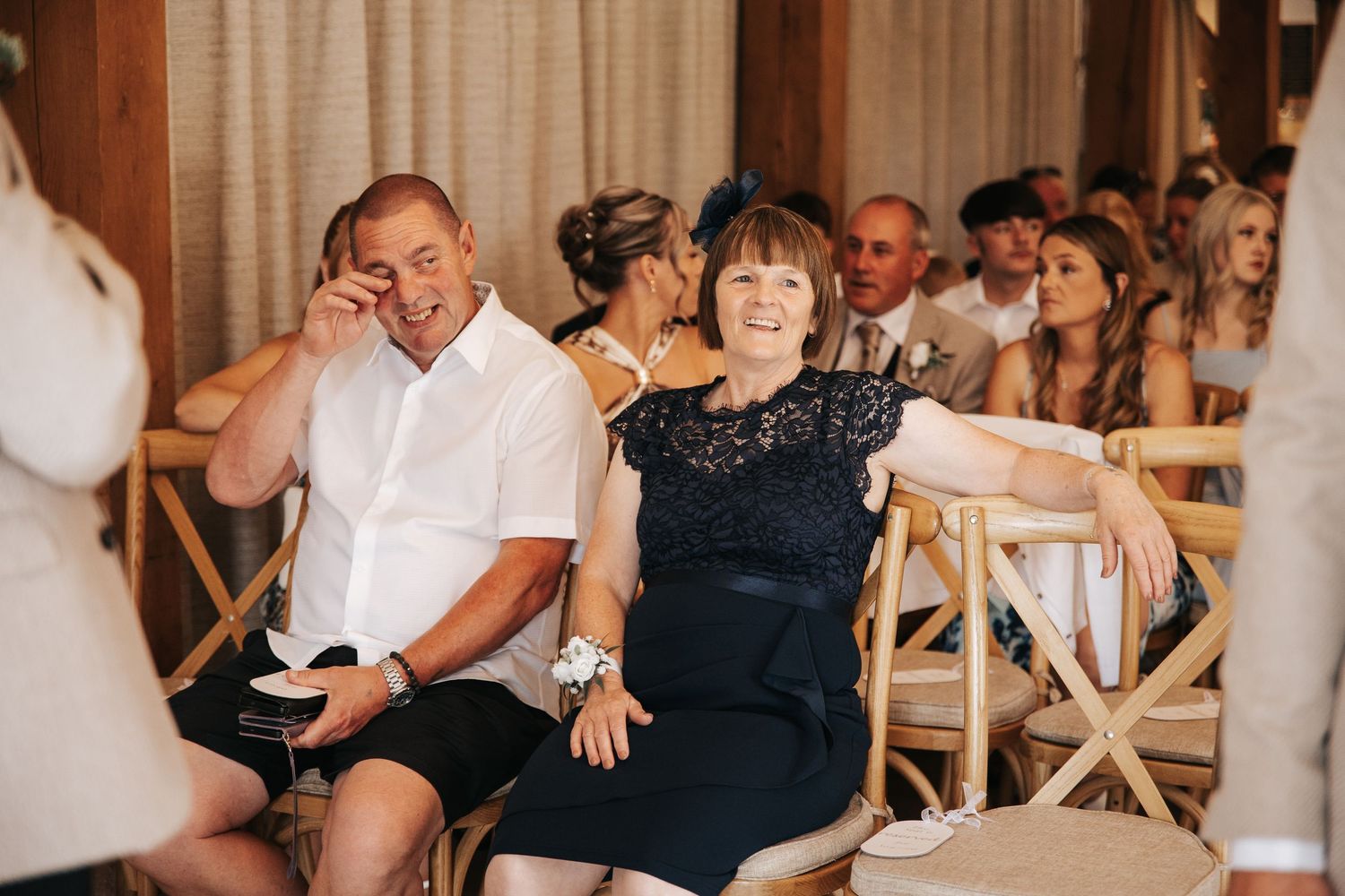 Wedding guests smiling and laughing during an outdoor ceremony, woman in navy lace dress seated in bamboo chairs.