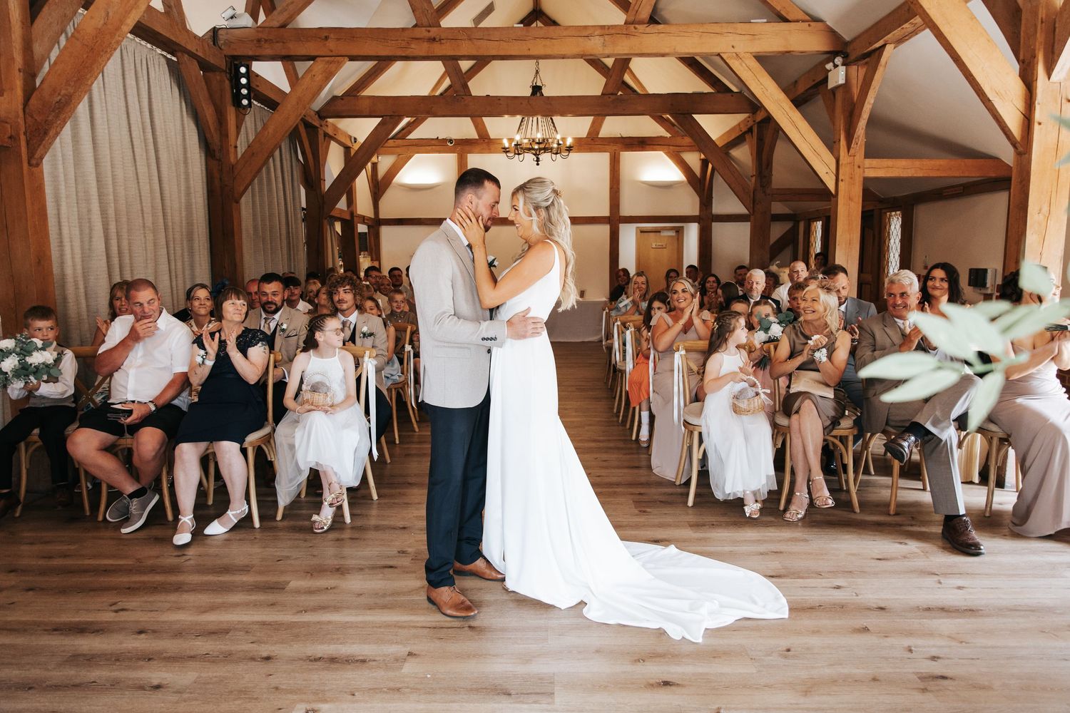 Bride and groom share first dance in rustic barn wedding venue with wooden beams and seated guests applauding.