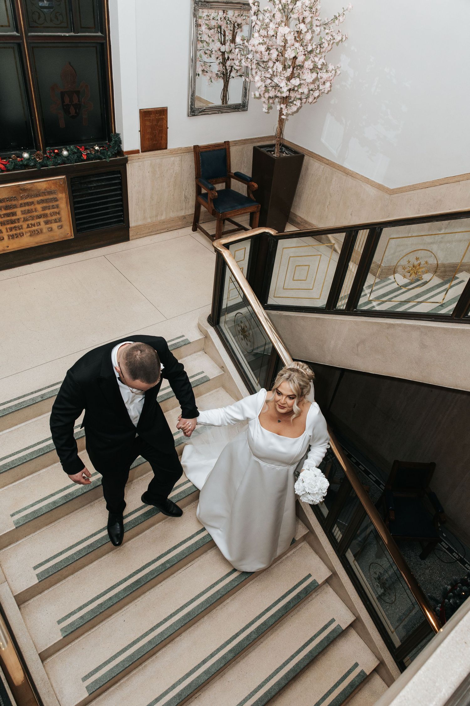 A couple walks down elegant stairs with decorative railings and a blooming cherry blossom tree.