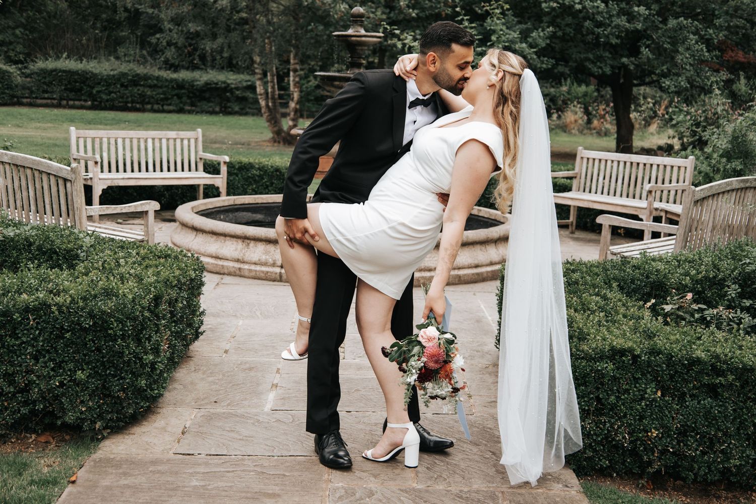 Wedding couple poses in garden setting with decorative benches and greenery.
