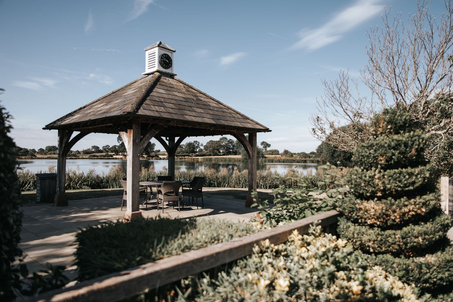 Wooden gazebo with dark roof beside a serene lake, surrounded by lush gardens under a blue sky.