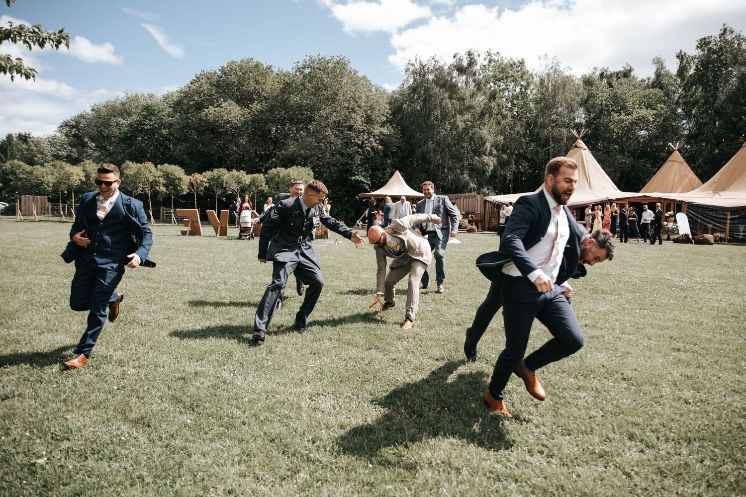 Groomsmen racing across green lawn at outdoor wedding venue with marquee tents and trees in background.