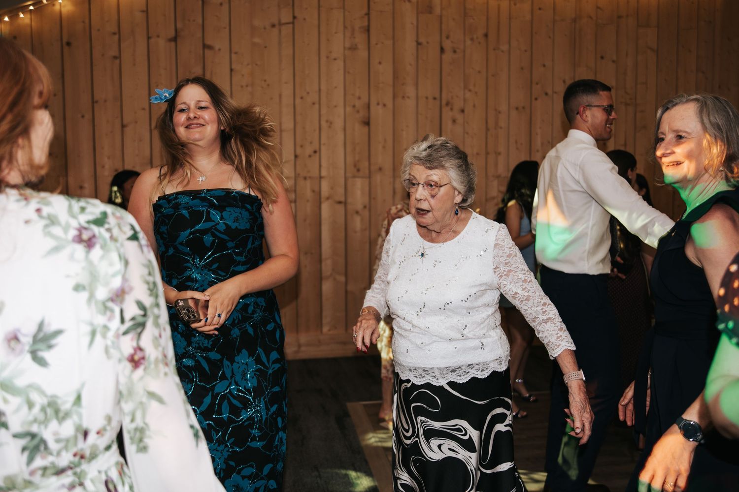 Women of different ages dancing joyfully at a wedding reception in a rustic wooden venue.