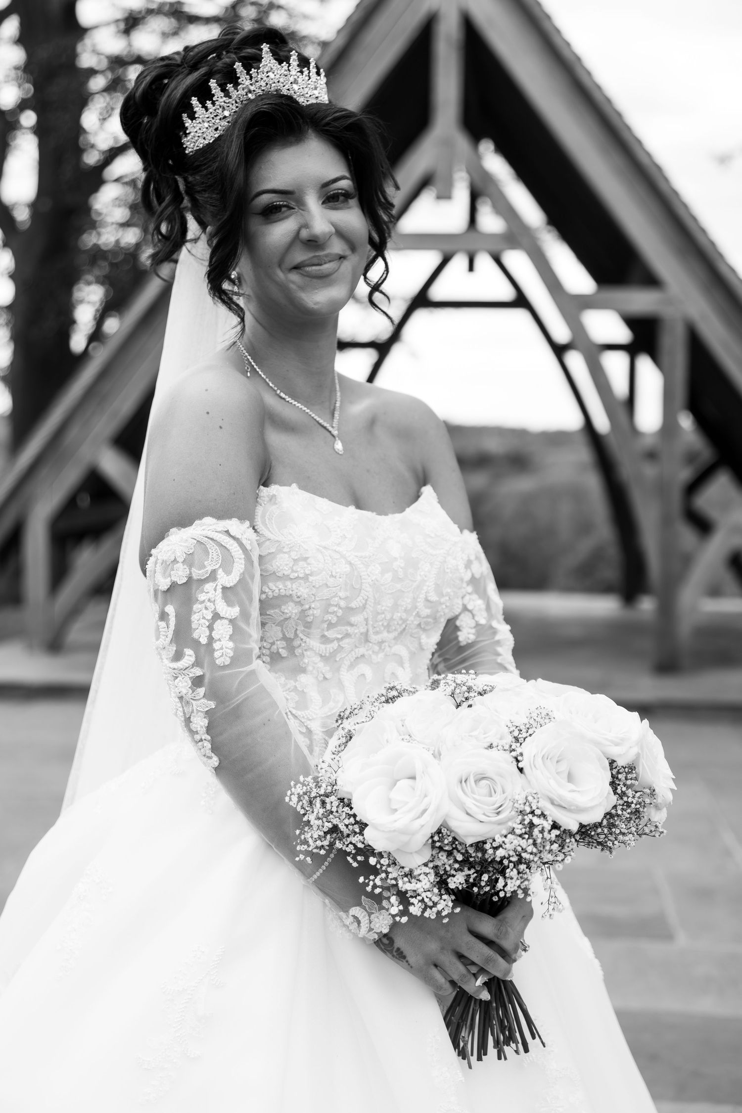Smiling bride in strapless lace gown and tiara holds white rose bouquet outdoors in black and white photo.