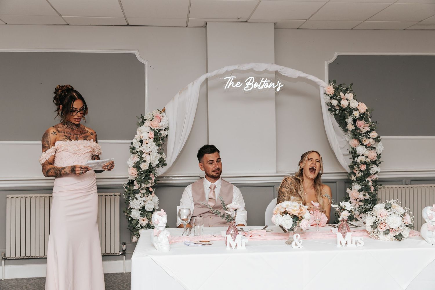 Bride and groom sit at wedding reception table with floral arch, as bridesmaid reads speech in elegant blush pink venue.