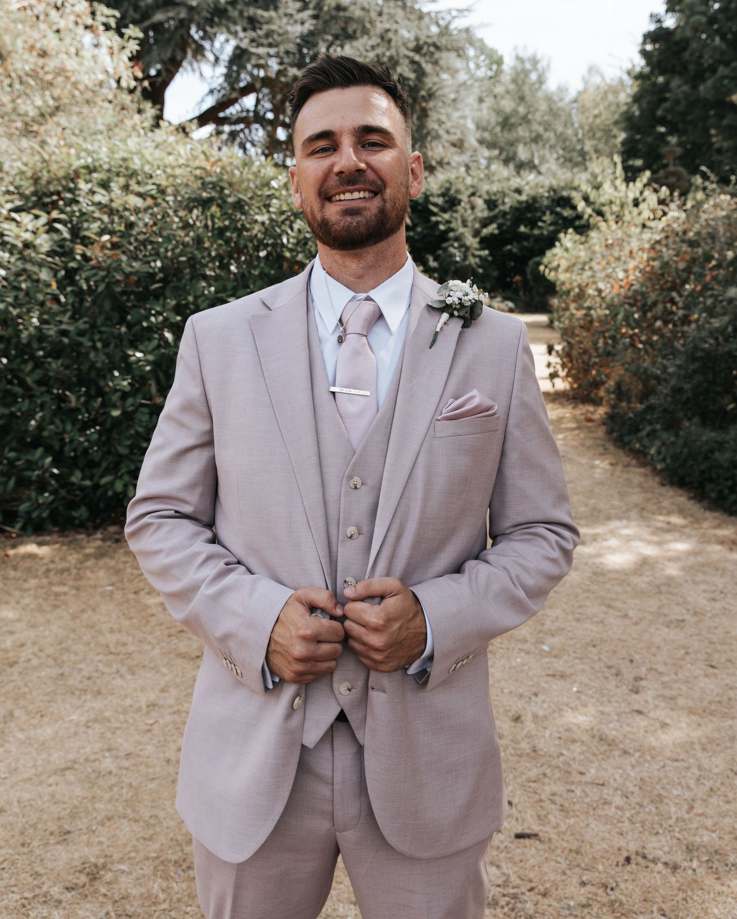Smiling groom in light gray three-piece suit with floral boutonniere posing outdoors among green hedges.