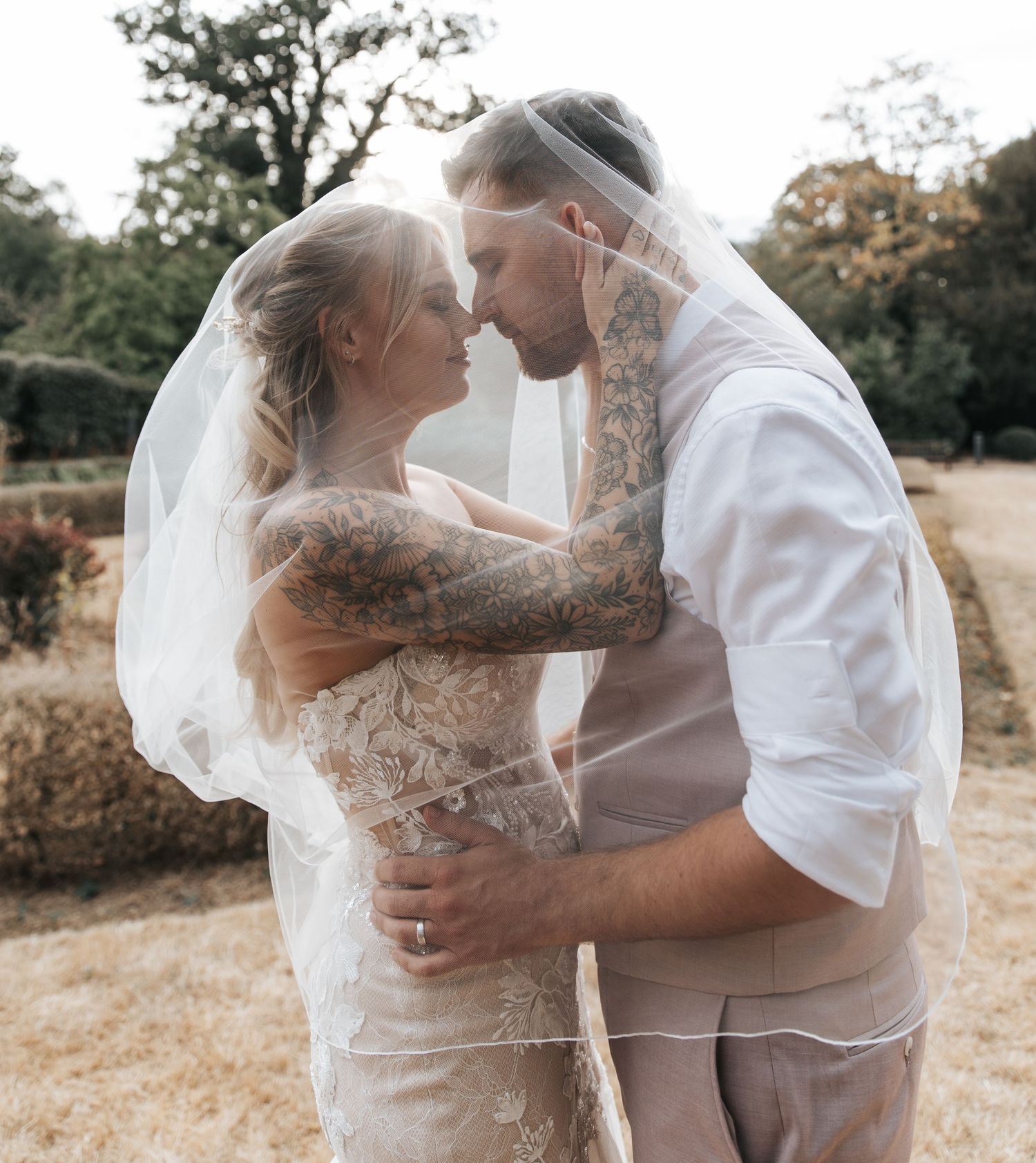 Bride and groom sharing romantic moment under wedding veil outdoors, groom with tattoos in white shirt, bride in lace gown.