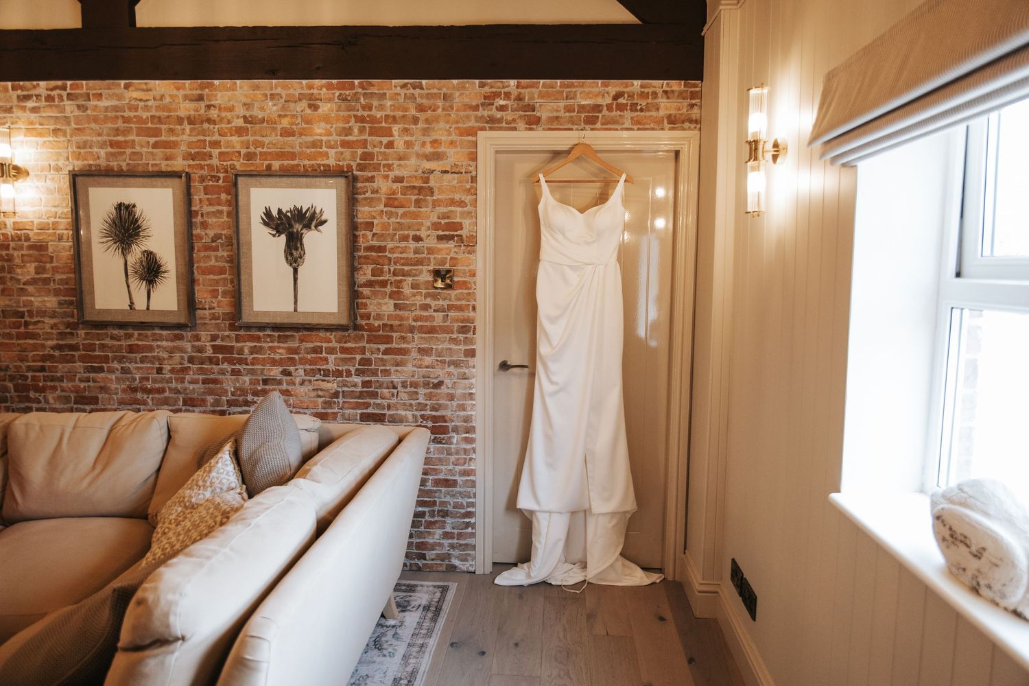 A white wedding dress hanging near a brick wall in a cozy bridal suite with a sofa and framed botanical art.
