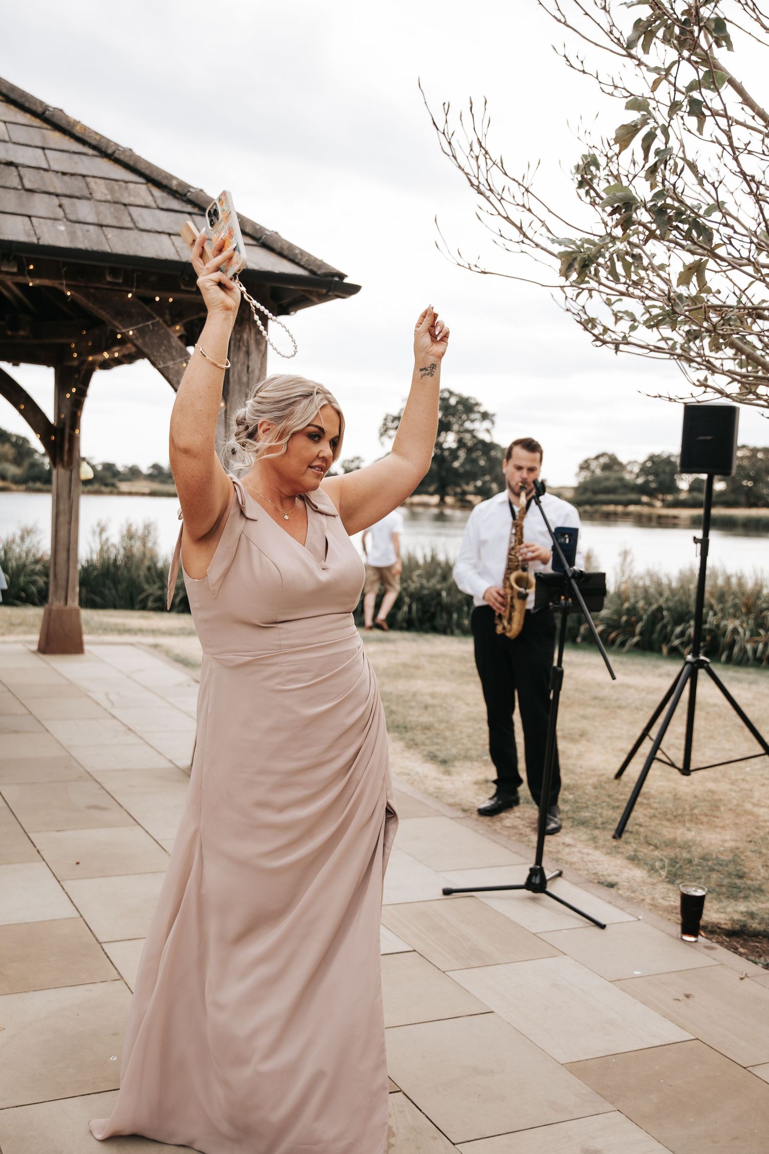 Woman in blush gown dancing at outdoor wedding reception while saxophonist performs near waterfront pavilion.