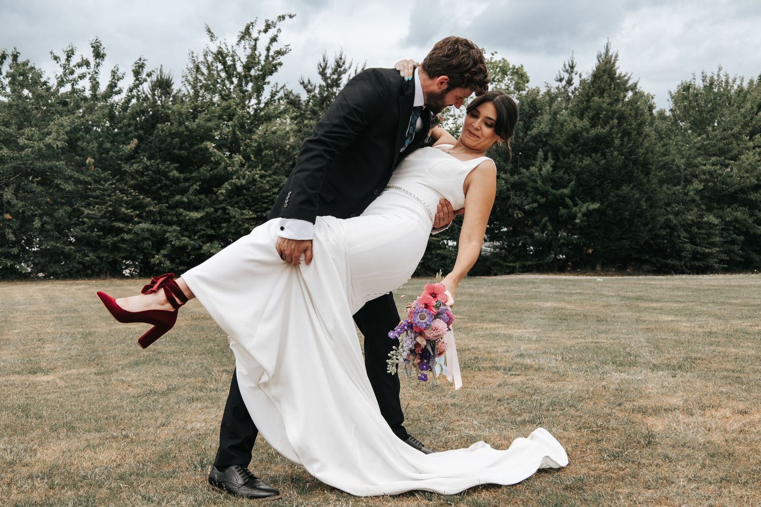 Wedding couple poses together on grassy field with cloudy sky backdrop.