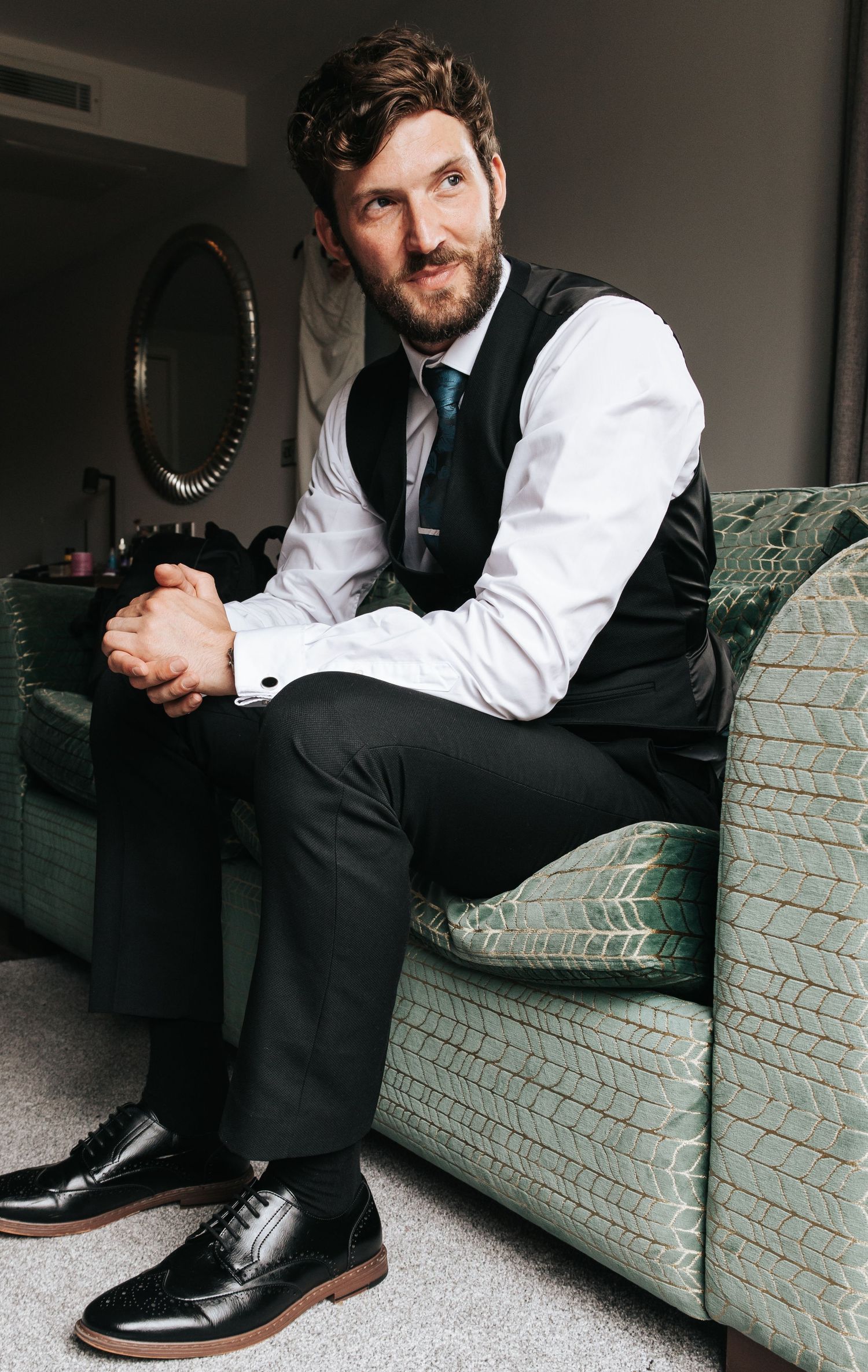 Man in formal attire relaxing on mint green couch during wedding preparation.