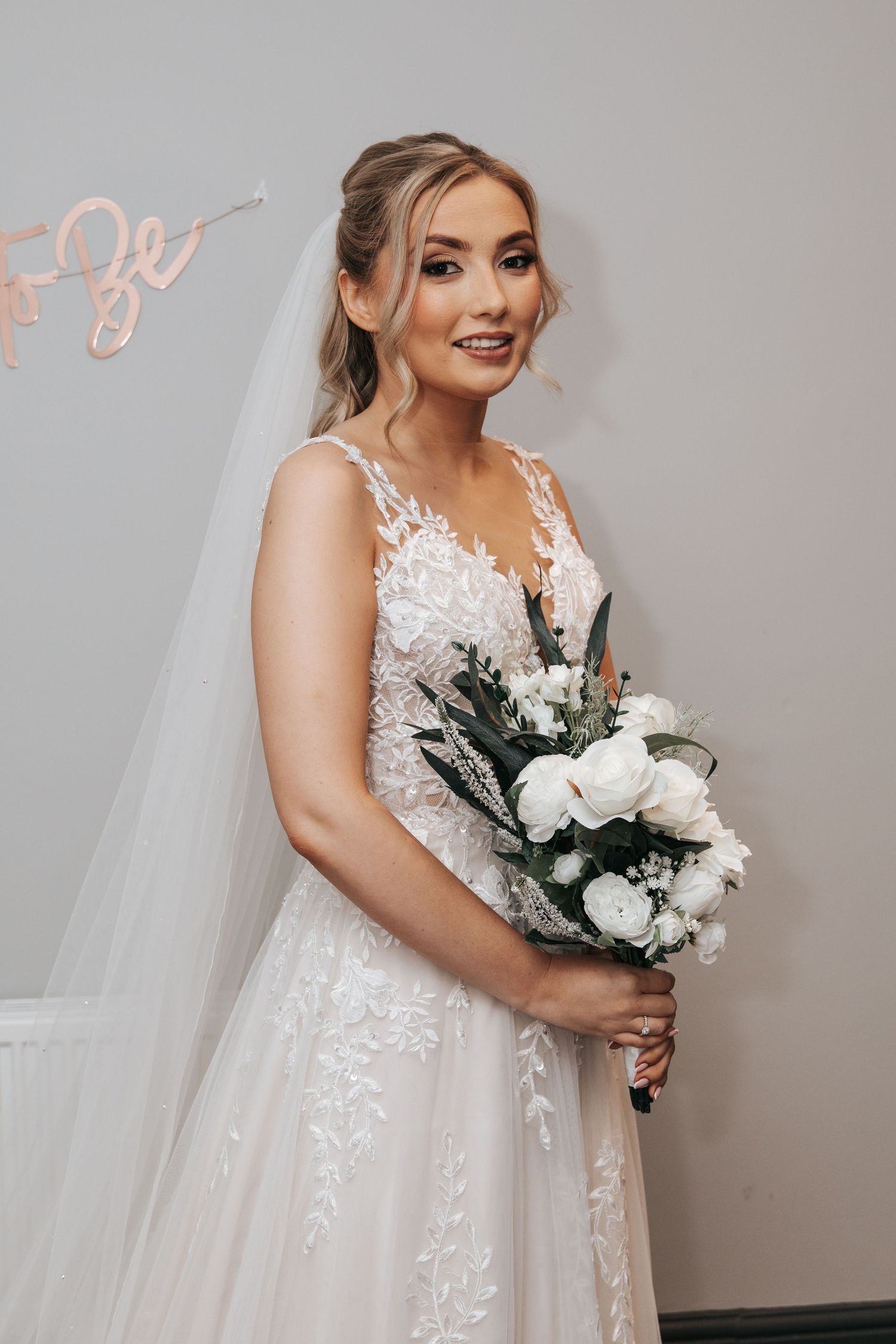 Bride in lace wedding gown holds elegant white and green bouquet against light background.
