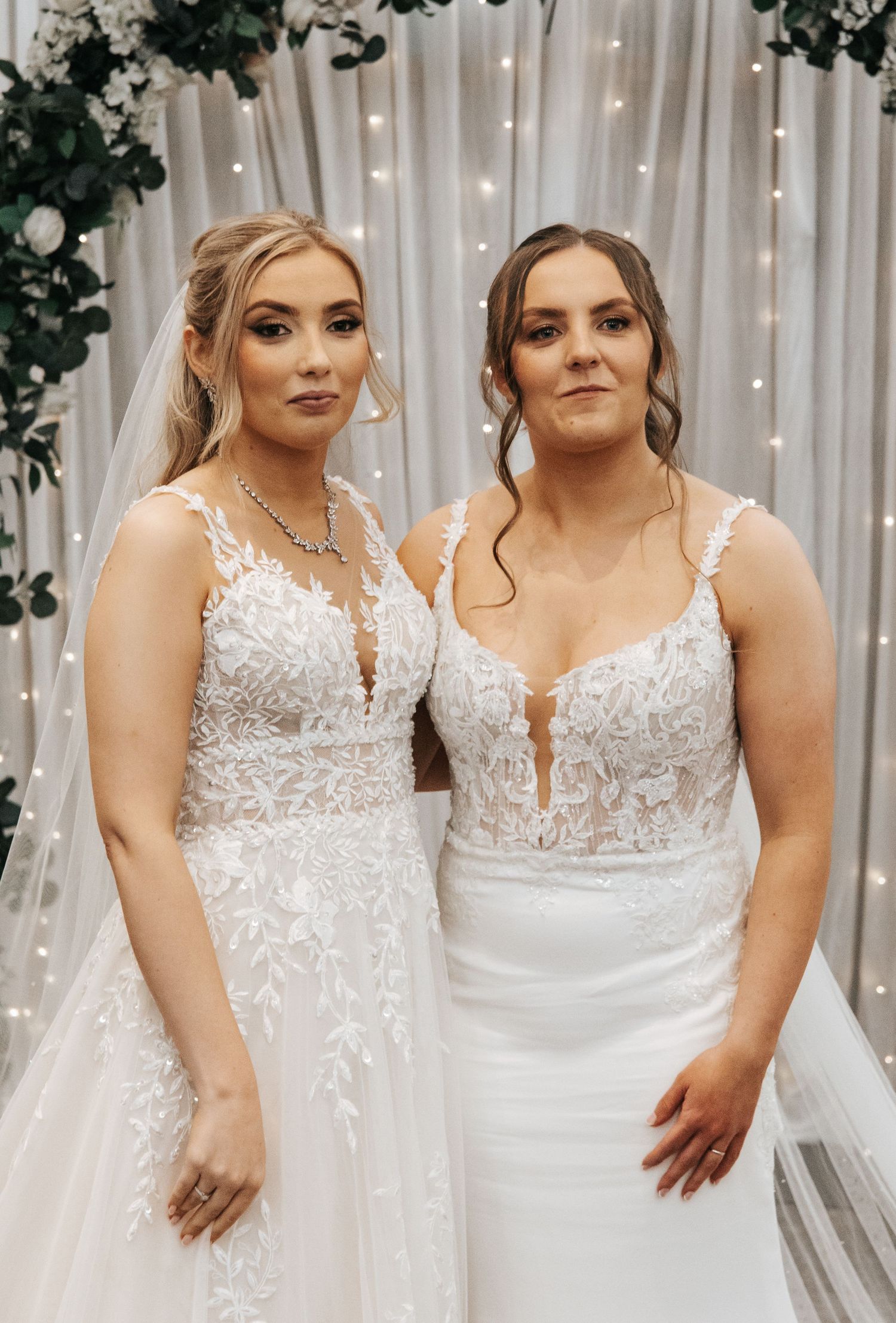 Two brides in detailed lace gowns stand close together against fairy light backdrop.