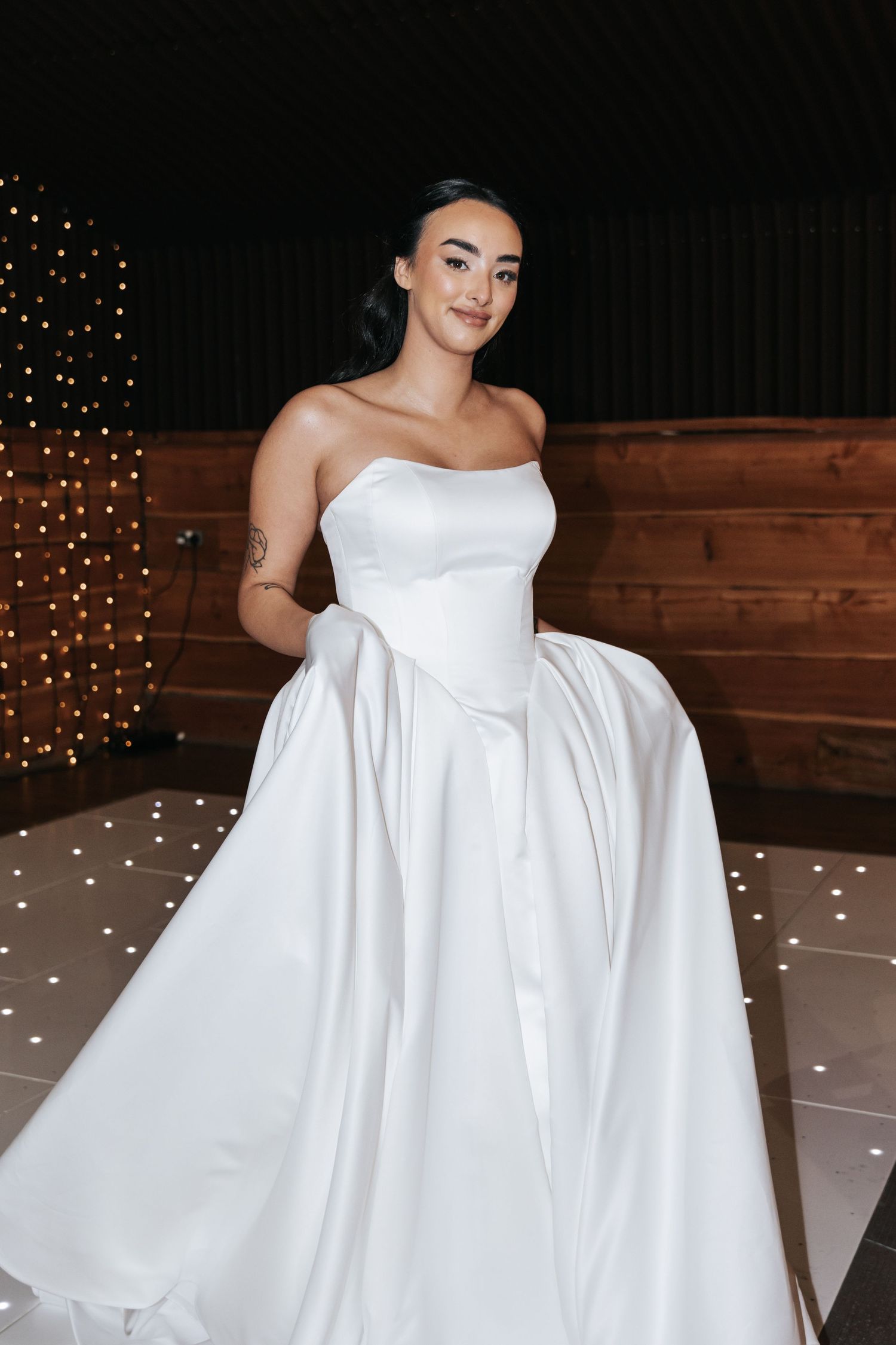 A bride in a white strapless ball gown poses in front of a rustic wood wall decorated with twinkling lights.