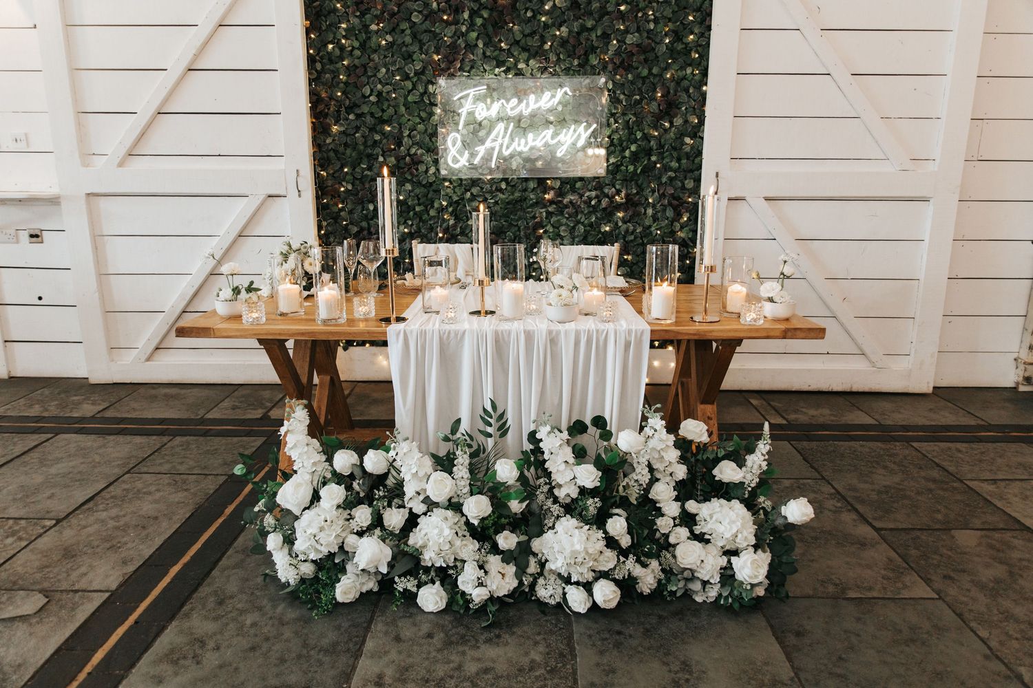 Elegant wedding sweetheart table with white floral arrangements, greenery wall backdrop, and glowing Forever & Always neon sign.