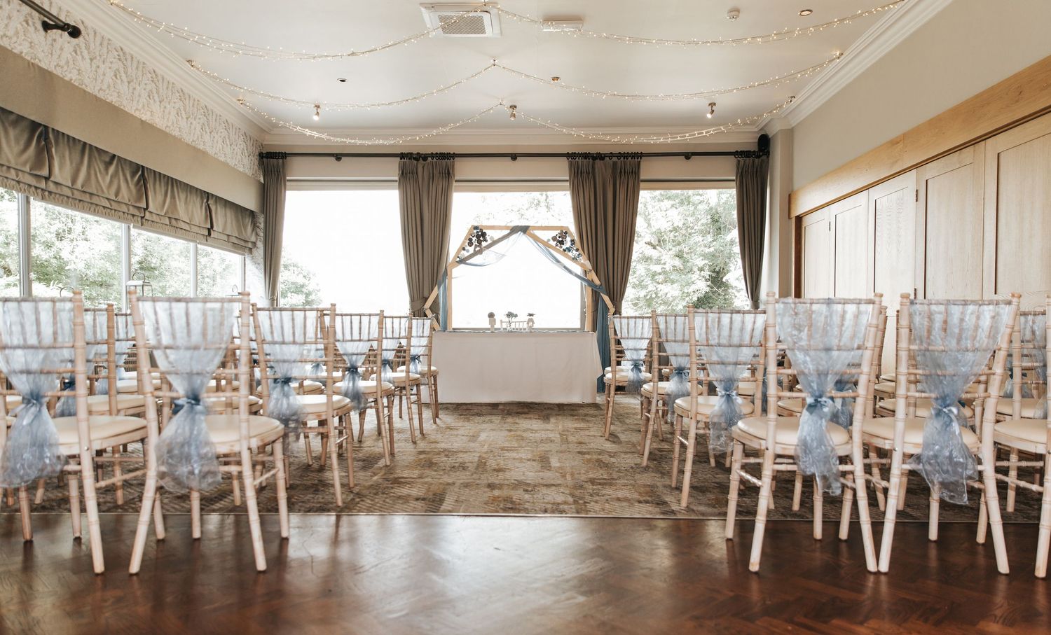 Elegant wedding ceremony room with white Chiavari chairs, blue sashes, floral arch, and large windows in a classic venue.