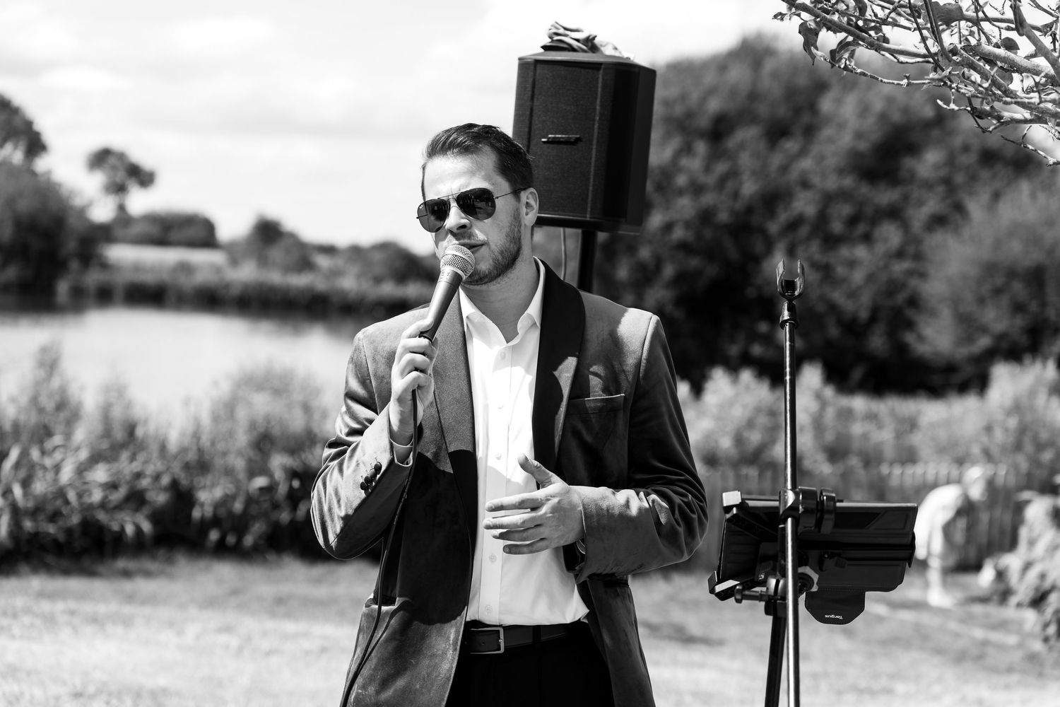 Man in suit and sunglasses singing into microphone outdoors at a garden event in black and white photo.
