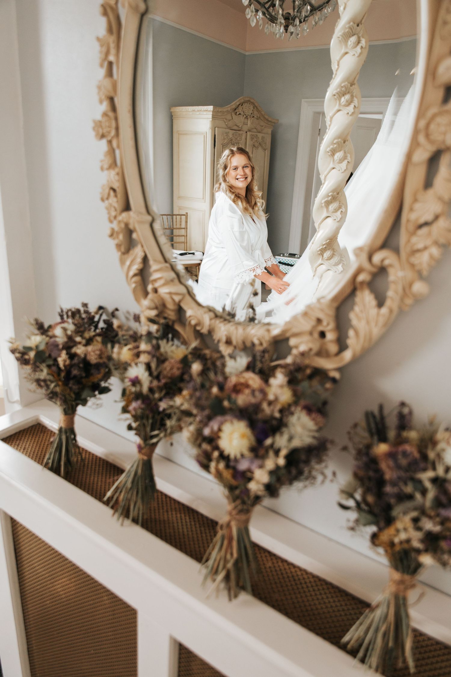 Bride in white robe smiling in ornate gold mirror, with dried floral bouquets arranged on a radiator cover.