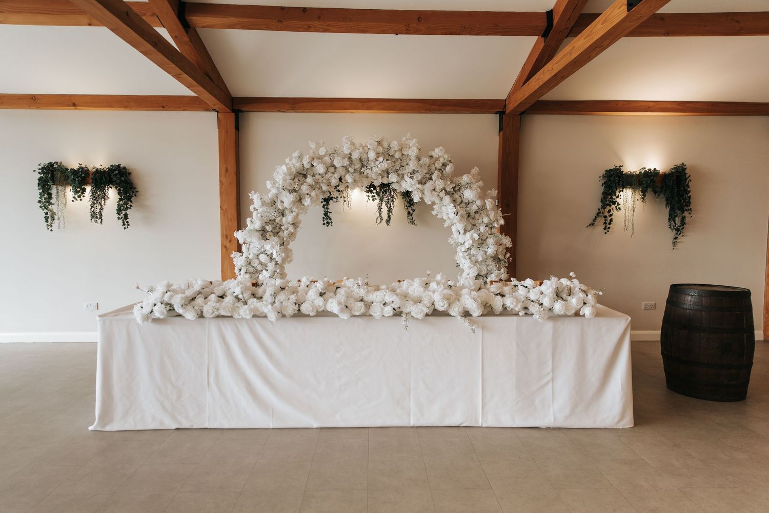Elegant wedding head table with white floral arch backdrop and draped tablecloth in a rustic venue.
