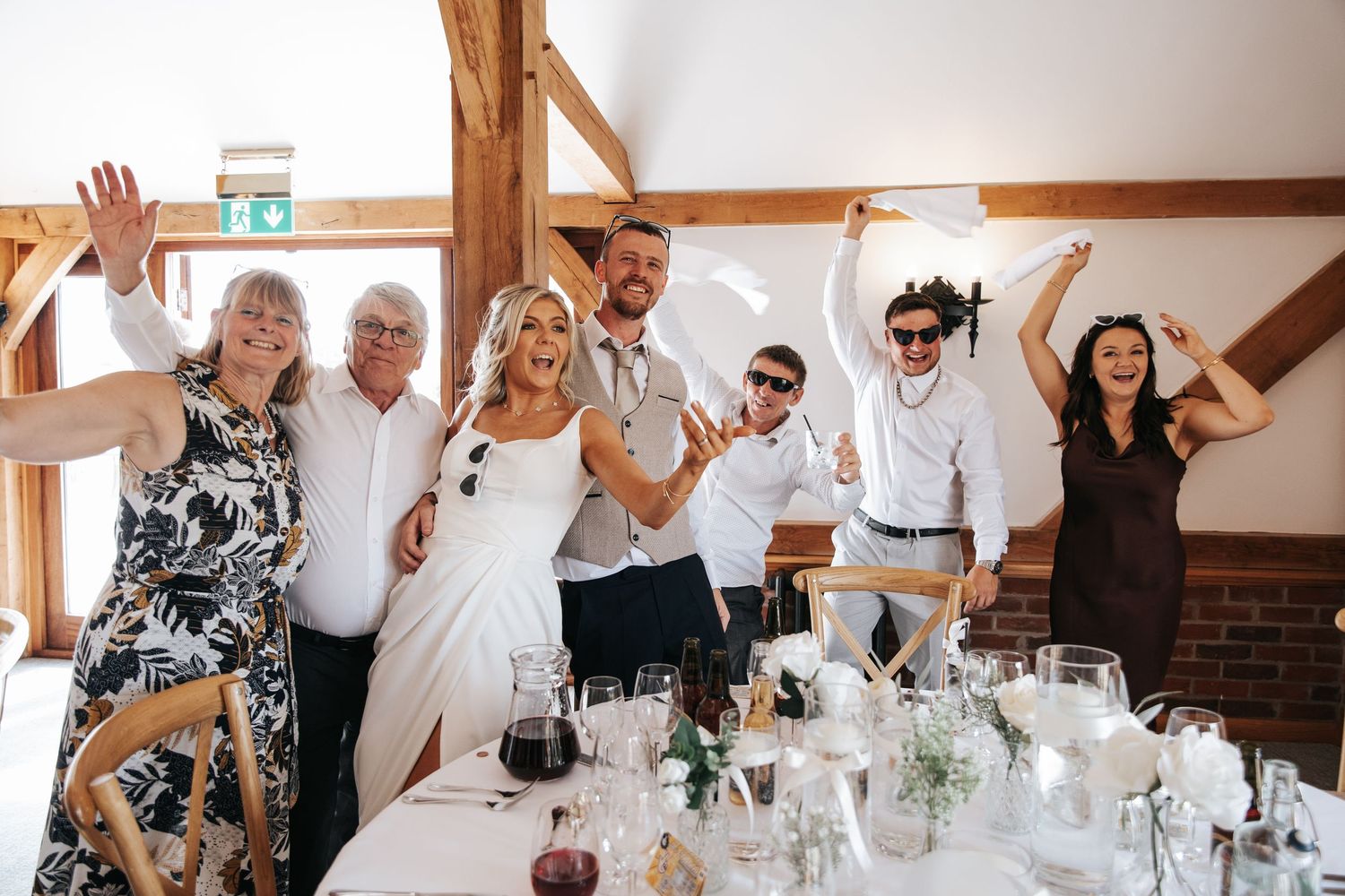 Wedding party celebrating at reception table, guests raising hands joyfully with bride in white dress centerpiece.