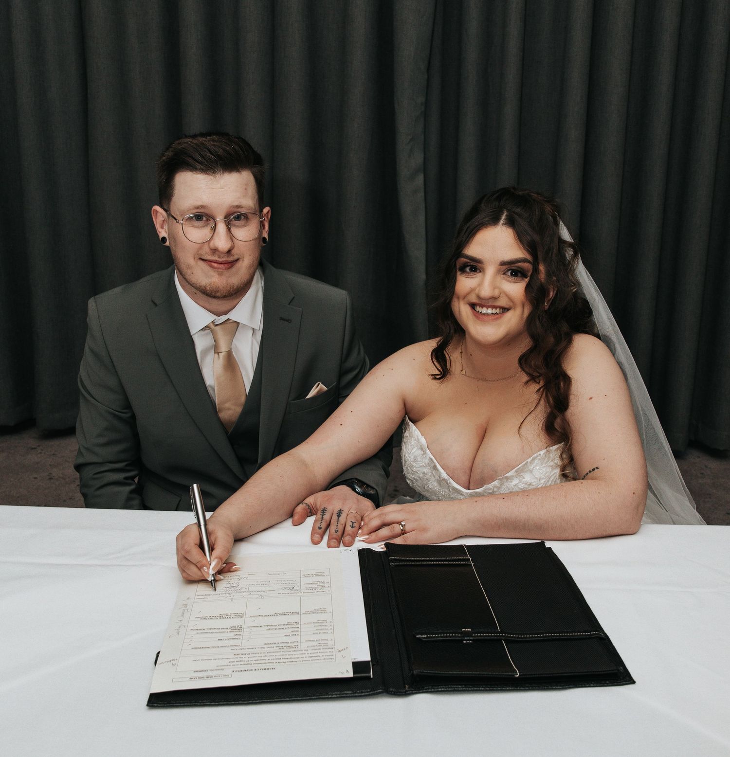 Wedding couple signs marriage certificate at formal ceremony with dark backdrop.