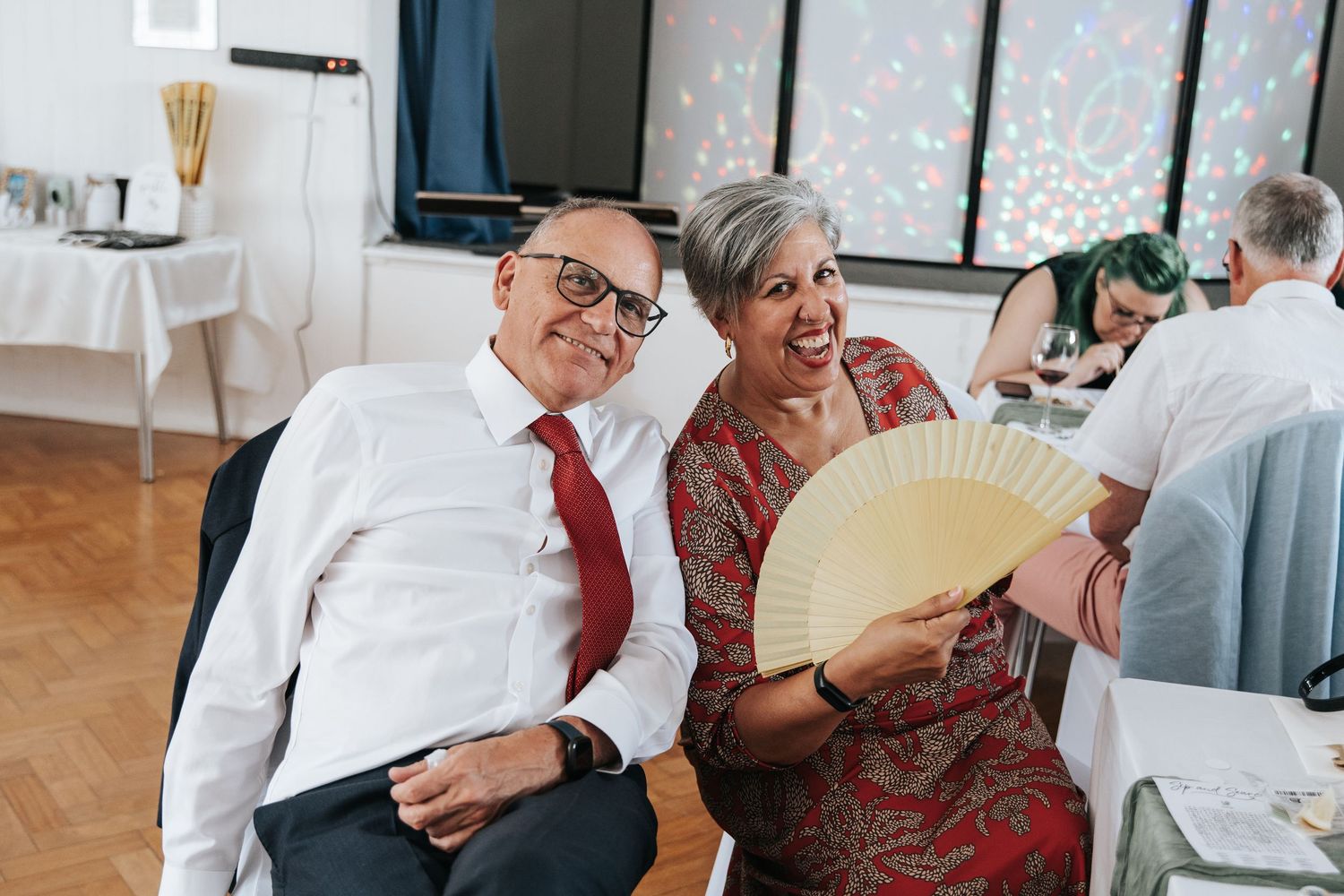 Elderly couple laughing together at a wedding reception, man in white shirt and red tie, woman holding a decorative fan.