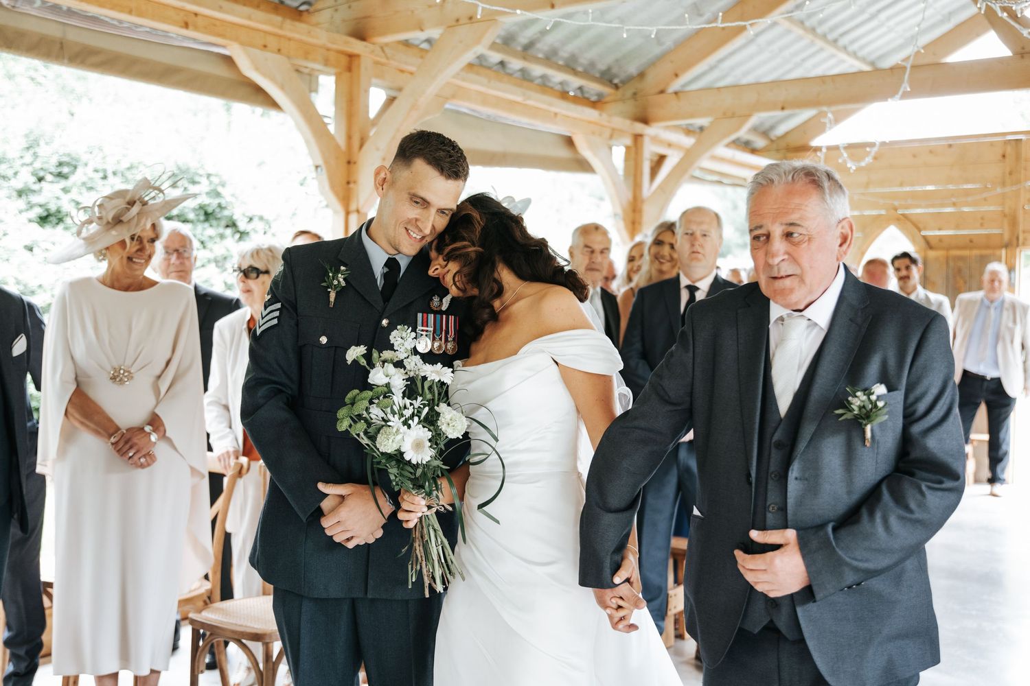 Military groom in uniform shares tender moment with bride holding bouquet as father walks her down the aisle.