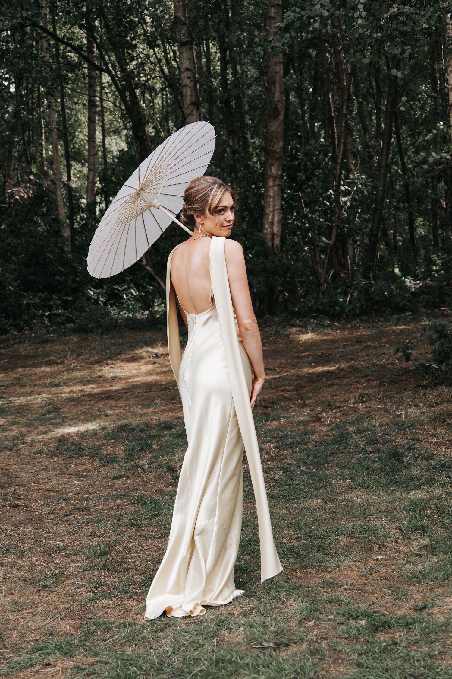 A bridesmaid in an ivory satin dress poses playfully with a white paper parasol in a forest setting.