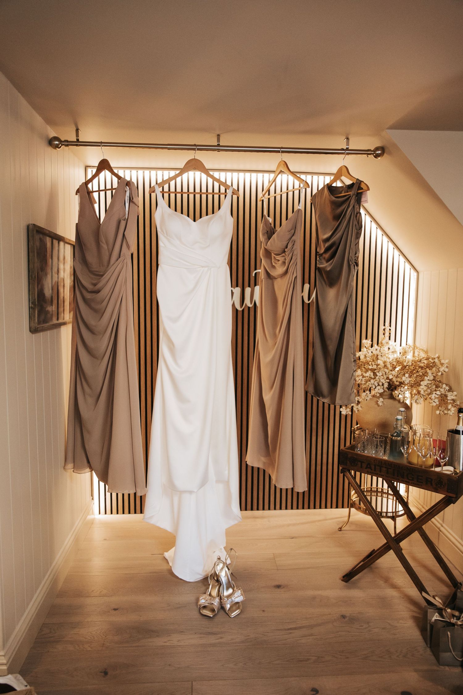 White wedding gown and taupe bridesmaid dresses hanging on a wooden rack in a warmly lit bridal suite.