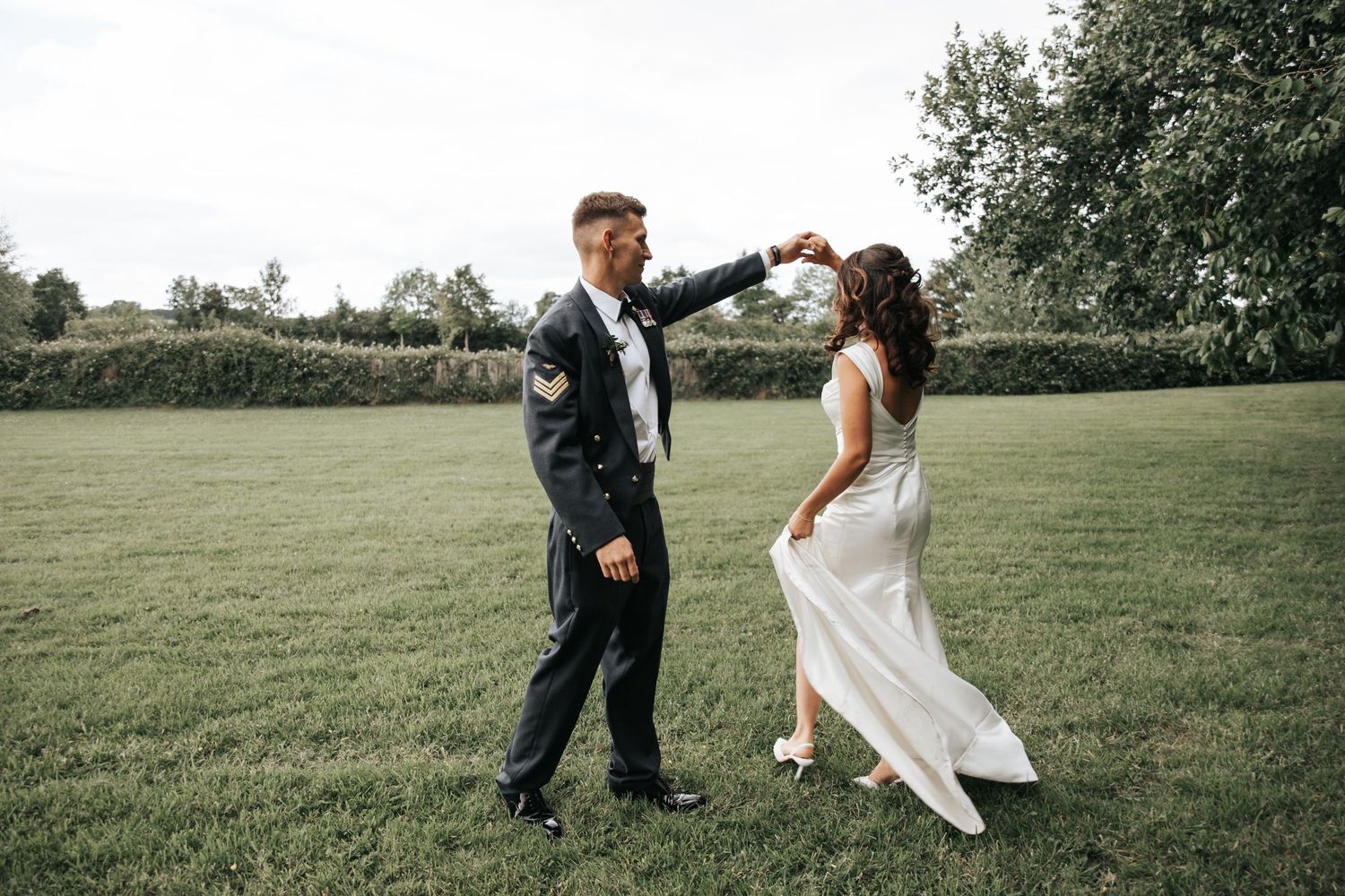 A groom in a dark suit twirls his bride in a white dress on a lush green lawn during an outdoor wedding.