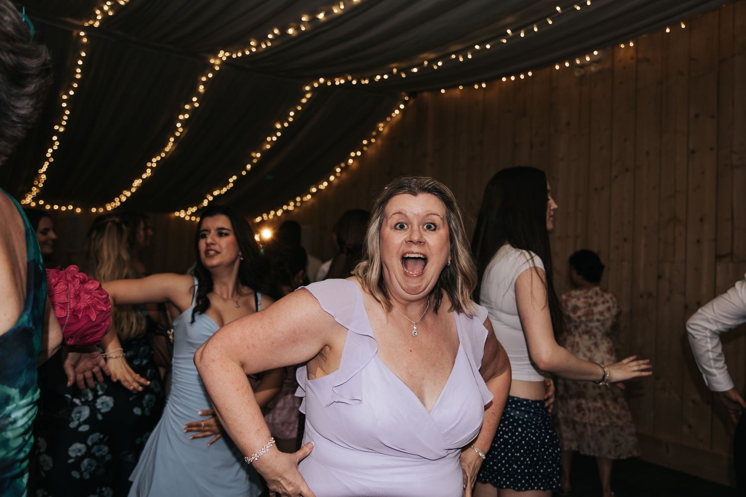 Excited woman dancing at a wedding reception under string lights in a rustic barn venue.