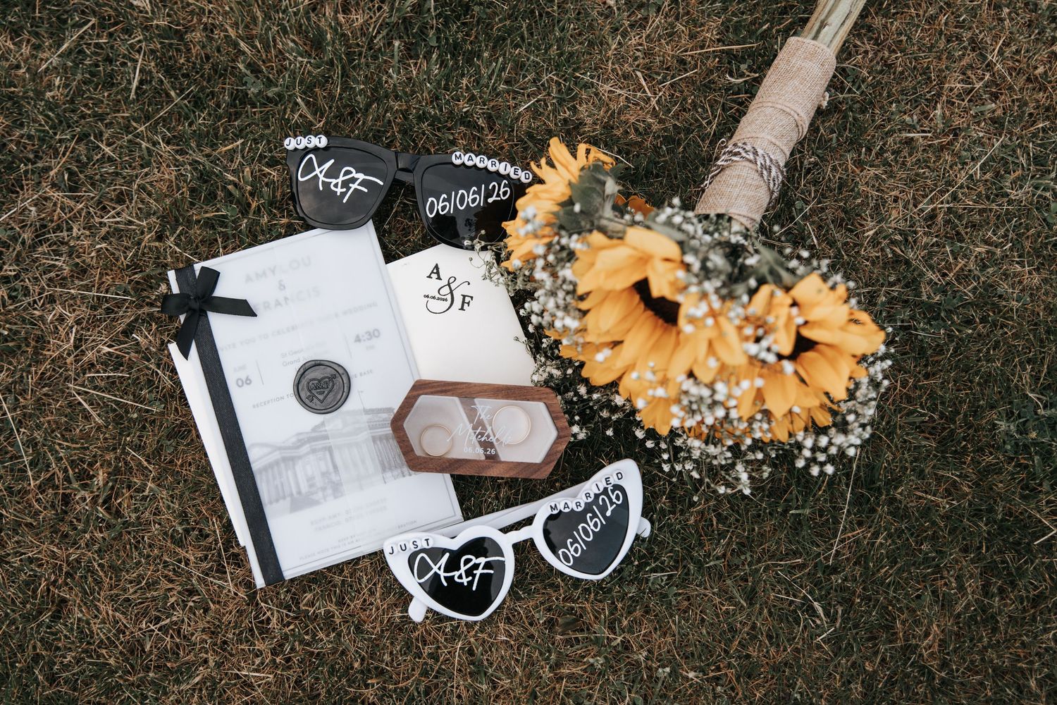 Wedding flat lay with sunflower bouquet, ring box, vow booklet, and custom sunglasses on grass.